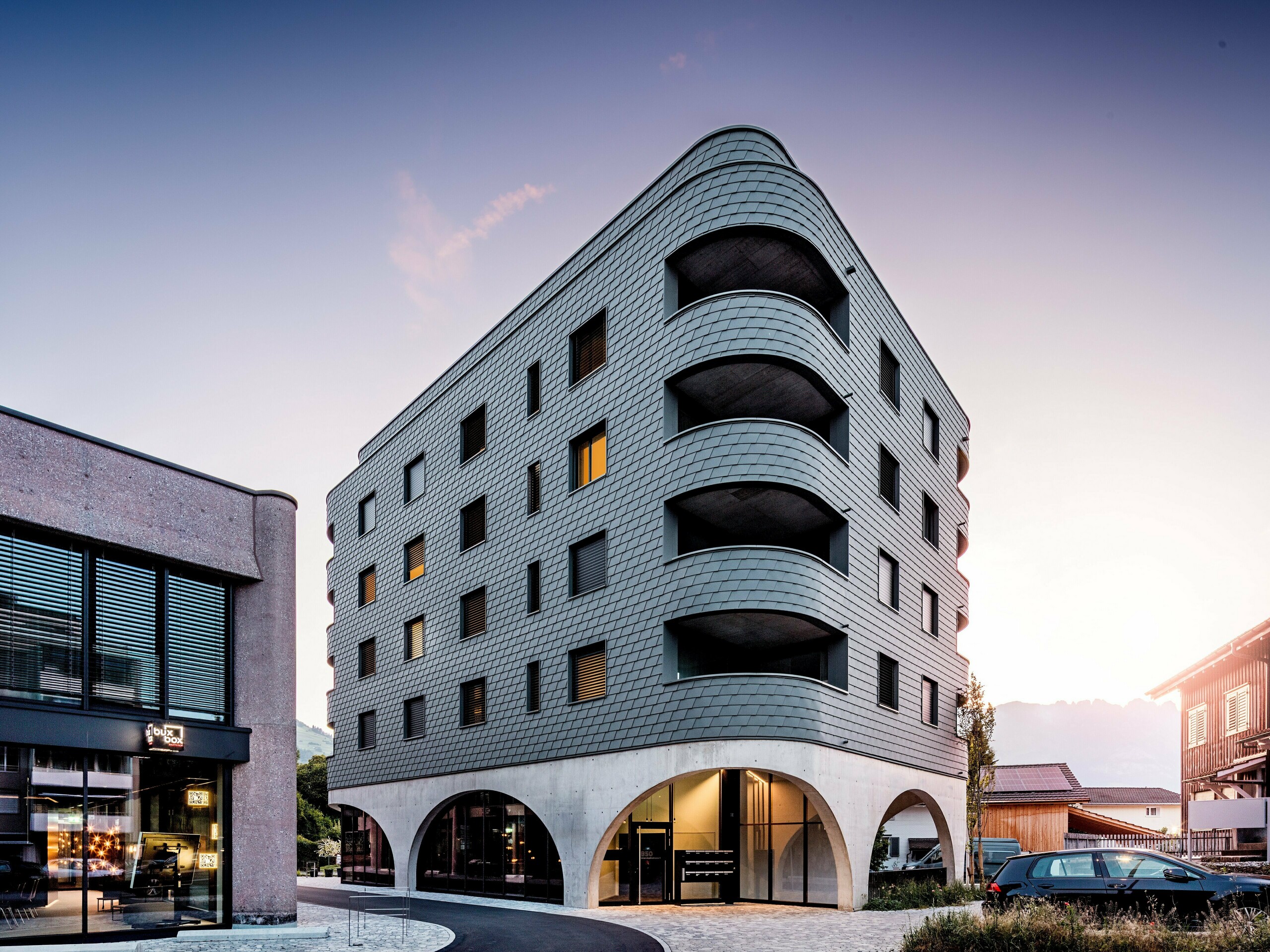 Moderní víceúčelová budova v Buchsu s výrazným, zaobleným, trojúhelníkovým tvarem a souvislou fasádou z PREFA fasádních šindelů v barvě P.10 světle šedé. Šupinovitý hliníkový plášť obaluje budovu jako plazí struktura a zdůrazňuje plynulé křivky a hluboce zapuštěné balkony. Nad masivním základem s elegantními betonovými arkádami působí obzvláště výrazně lehká a povětrnostním vlivům odolná fasáda PREFA. Precizně položené hliníkové šindele zajišťují homogenní, odolný a nenáročný plášť budovy s vysokou rozpoznatelností.