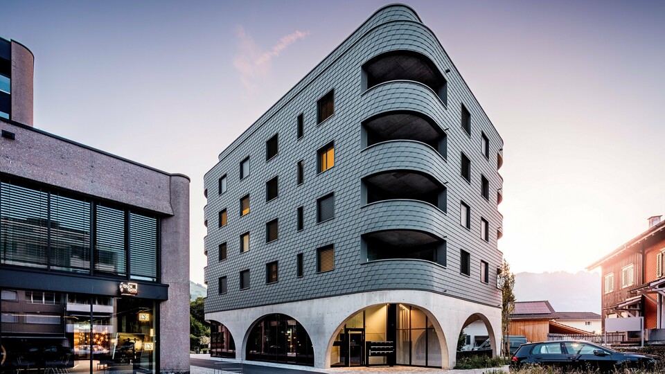 Moderní víceúčelová budova v Buchsu s výrazným, zaobleným, trojúhelníkovým tvarem a souvislou fasádou z PREFA fasádních šindelů v barvě P.10 světle šedé. Šupinovitý hliníkový plášť obaluje budovu jako plazí struktura a zdůrazňuje plynulé křivky a hluboce zapuštěné balkony. Nad masivním základem s elegantními betonovými arkádami působí obzvláště výrazně lehká a povětrnostním vlivům odolná fasáda PREFA. Precizně položené hliníkové šindele zajišťují homogenní, odolný a nenáročný plášť budovy s vysokou rozpoznatelností.