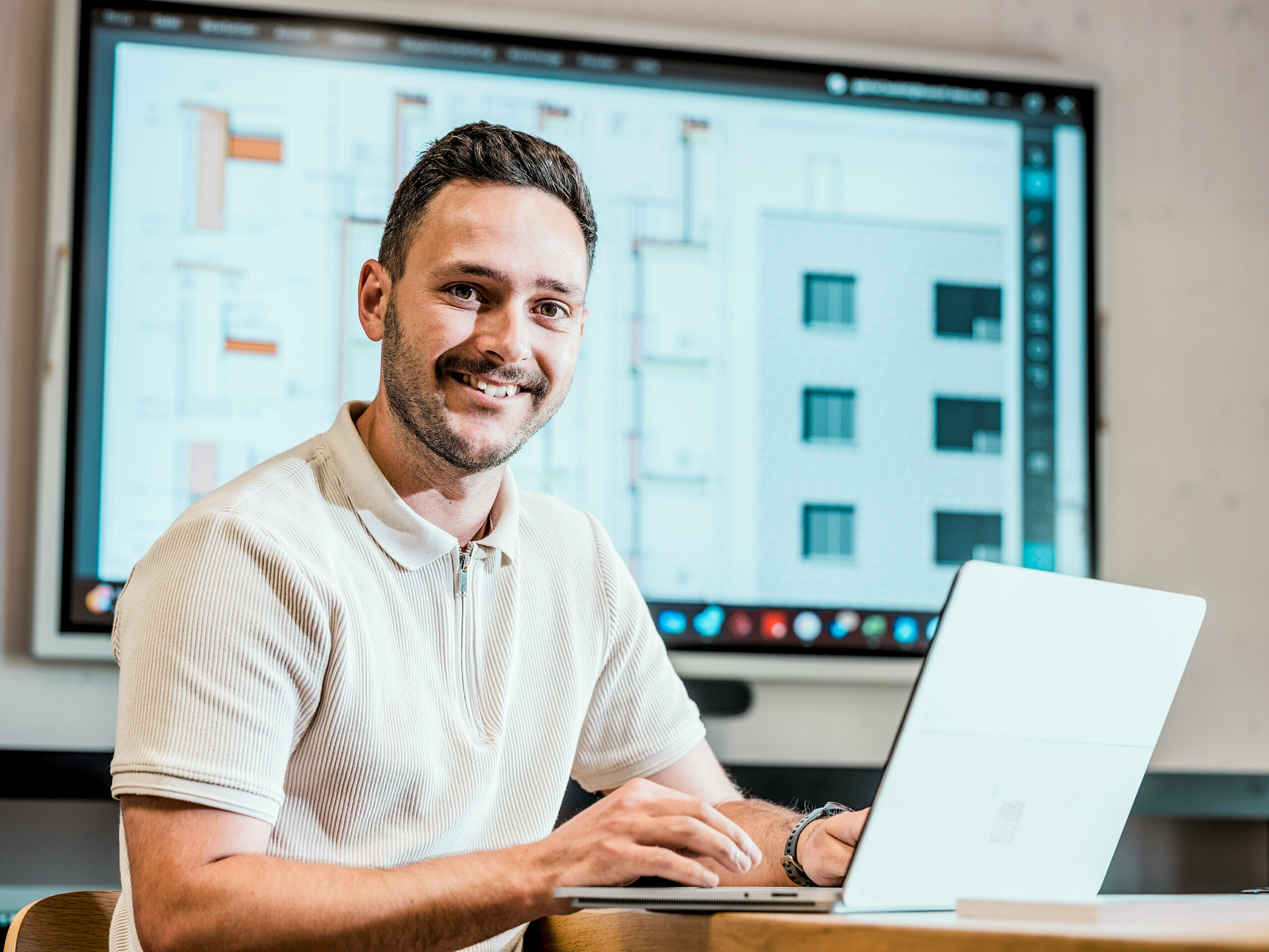 Portrét Gabriela Landolta, zaměstnance společnosti Künzli Davos AG, u svého notebooku před velkou obrazovkou zobrazující digitální stavební plány a detaily fasády. Monitor zobrazuje technické výkresy a pohledy moderní budovy s měřítkovou fasádou, postavené s použitím fasádních šindelů PREFA v barvě P.10 světle šedá. Inovativní hliníková fasáda přesvědčí svou nízkou hmotností, odolností vůči povětrnostním vlivům a dlouhou životností. Precizní plánování a použití vysoce kvalitních produktů PREFA tvoří základ pro rozměrově stabilní, nenáročný na údržbu a architektonicky atraktivní plášť budovy.