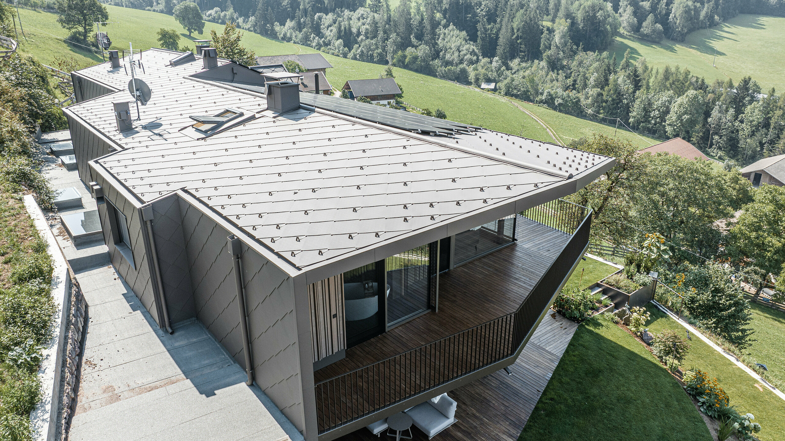 Pohled na zmodernizovaný dvojdům v Möltenu v Jižním Tyrolsku vybavený odolnými falcovanými a falcovanými šablonami 44x44 v barvě P.10 tmavě hnědé. Design střechy a fasády kombinuje robustní hliník s dřevěnými prvky, obklopený malebnou krajinou s rozsáhlými výhledy na zalesněné kopce. Redesign plynule integruje zrekonstruovanou stávající budovu a nově vybudovaný areál do harmonického celku se zaměřením na dřevo jako stavební materiál.