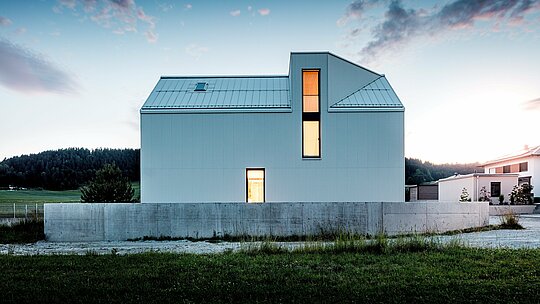 Obrázek ukazuje výrazný rodinný dům ve Freistadtu, který zaujme svými čistými liniemi a minimalistickou architekturou. Čistě bílá fasáda PREFALZ, vyrobená z ohýbaného hliníku, tvoří výraznou geometrickou strukturu, která budově dodává moderní a nadčasový vzhled. Strmá šikmá střecha s integrovaným vikýřem a velká okna zajišťují dostatek přirozeného světla, díky čemuž interiér působí otevřeně a příjemně. Použití hliníku PREFALZ zaručuje nejen estetickou stálost, ale také vynikající odolnost vůči povětrnostním vlivům a trvanlivost, díky čemuž je dům funkční i udržitelný. Jasný designový jazyk a stylové detaily přispívají k elegantnímu a zároveň praktickému řešení pro moderní rezidenční architekturu.