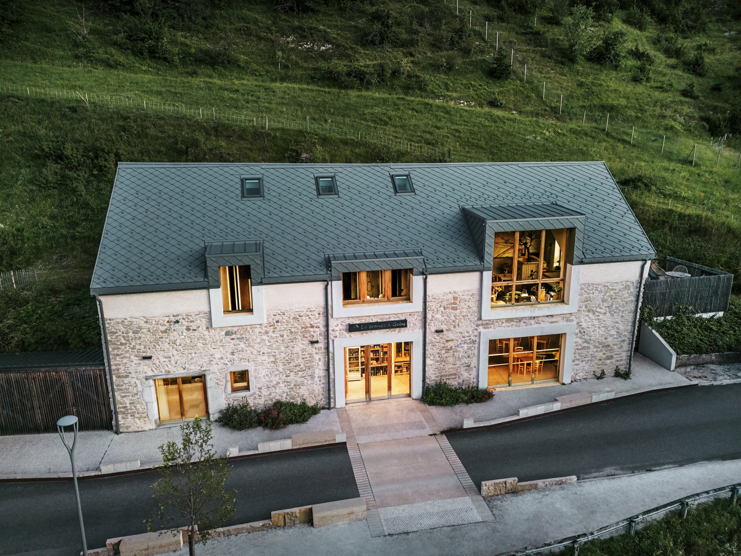 Večerní pohled na zrekonstruovanou stodolu "La Grange à Gaby" v Engins, Francie. Budova kombinuje tradiční kamenné zdivo s moderní hliníkovou střechou PREFA z falcovaných šablon 29 × 29 cm. Velká okna a prosvětlené interiéry podtrhují přívětivou atmosféru nového kulturního centra.