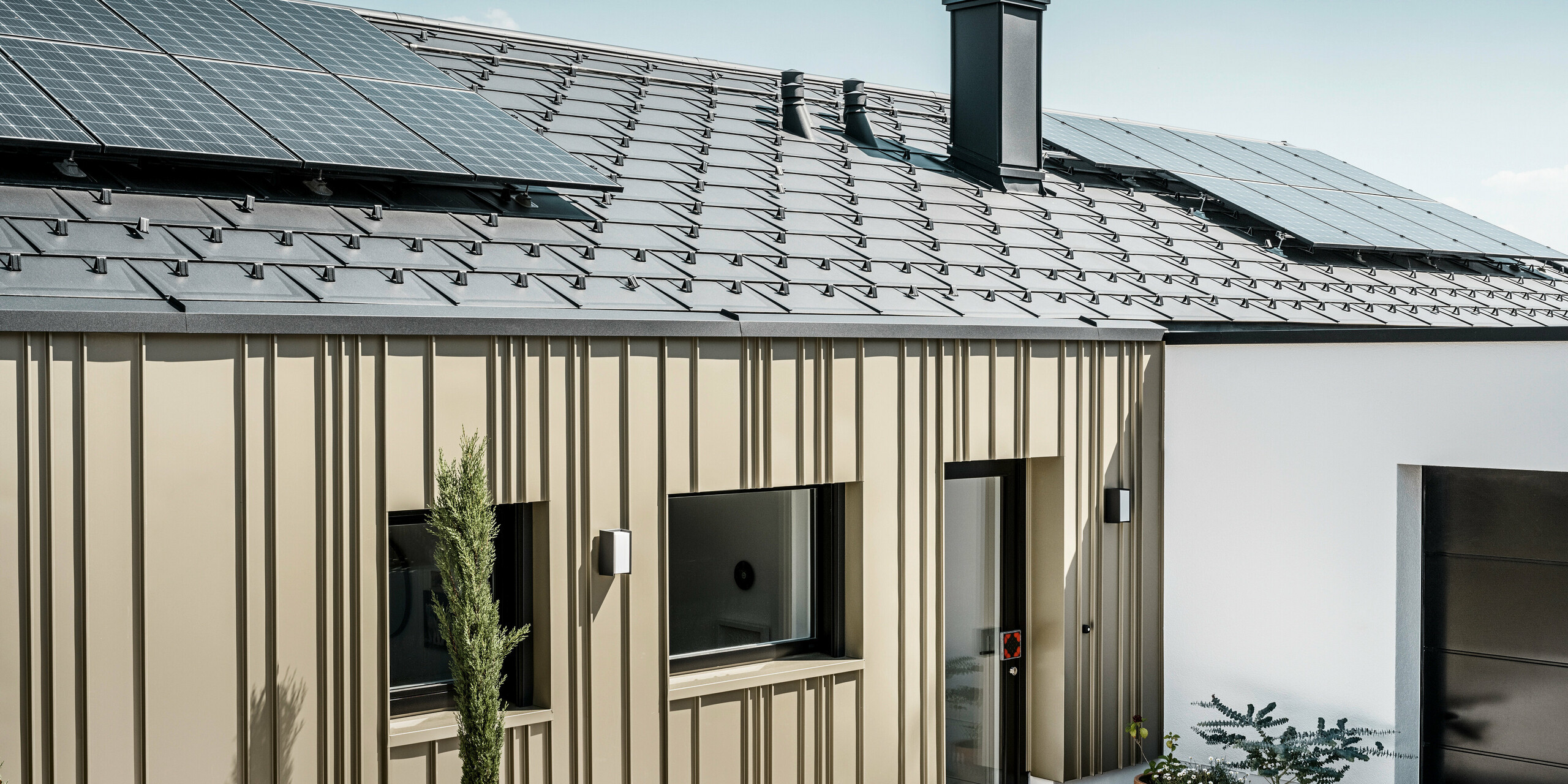 Energieeffizientes Einfamilienhaus mit einem langlebigen PREFA Aluminiumdach in Anthrazit, ausgestattet mit Photovoltaikmodulen. Die zukunftsweisende Wohnlösung wird zudem von einer robusten und widerstandsfähigen PREFA Aluminiumfassade in Lichtbronze betont. Die Fassade weist eine vertikale Strukturierung auf, die durch die anthrazitfarbene PREFA Dachplatte R.16 ergänzt wird. Diese Kombination bietet nicht nur ästhetische Schönheit, sondern auch Nachhaltigkeit und Energieunabhängigkeit in der modernen Architektur. Ergänzt wird der nachhaltige Bau durch eine PV-Anlage auf dem Dach.