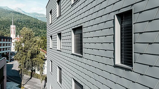 Detailní pohled na fasádu Bernerhausu v Buchsu s přesně položenými falcovanými šindeli PREFA v barvě P.10 světle šedé. Šupinatý hliníkový povrch vytváří jemný, zářivý vzor a zdůrazňuje jasně definované okenní otvory s hlubokými ostěními. Lehký, povětrnostním vlivům odolný hliník PREFA zajišťuje homogenní, rozměrově stabilní a nenáročný na údržbu plášť budovy. Na alpském pozadí se vysoce kvalitní kovová fasáda stává moderním poutavým prvkem s nadčasovým vzhledem.