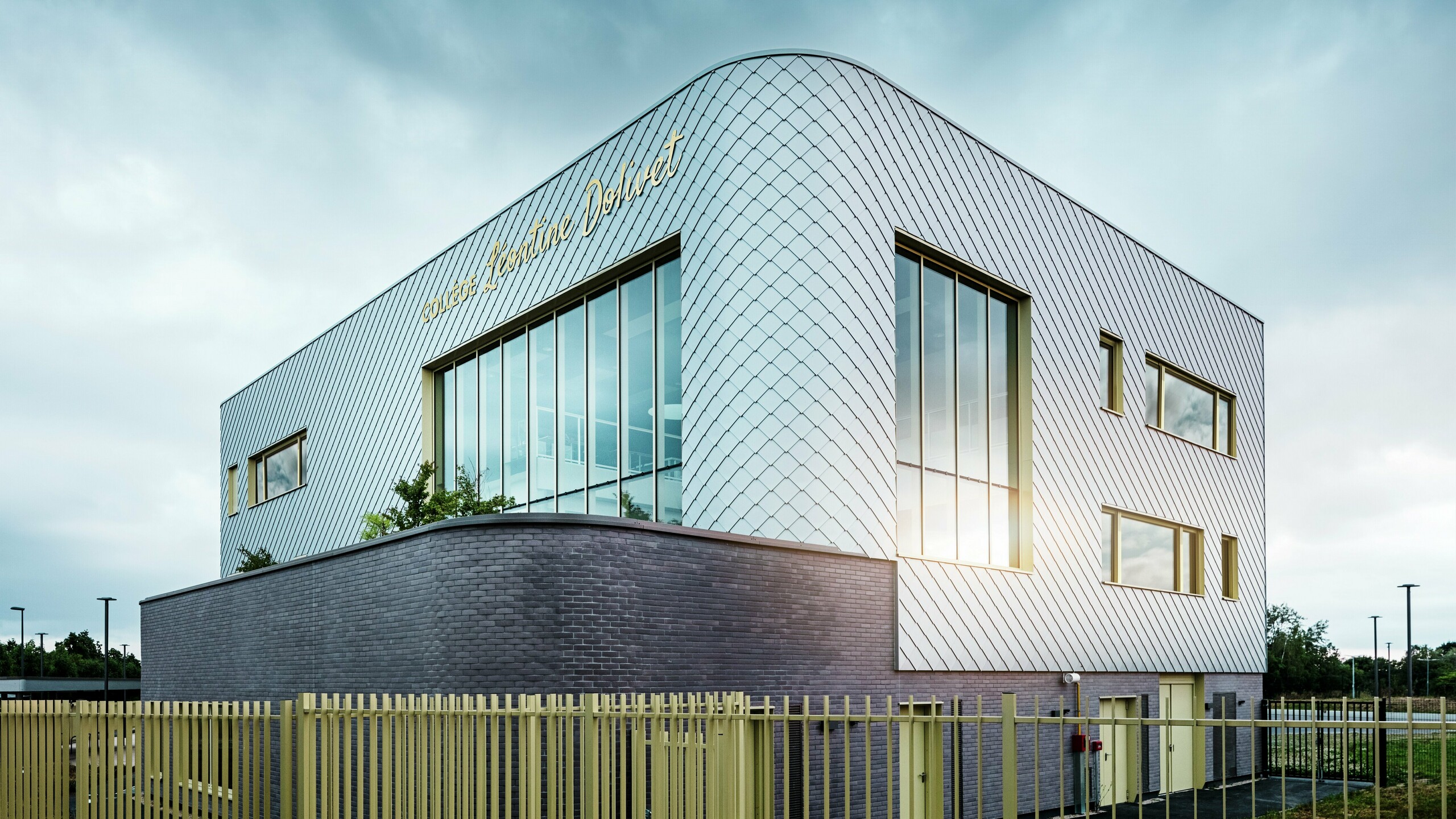 Das Foto zeigt eine markante Ecke des Collège Léontine Dolivet mit seiner geschwungenen Aluminiumfassade aus silbermetallicfarbenen PREFA-Wandelementen. Die großen Glasflächen reflektieren das Licht, während die gelben Fensterrahmen Akzente setzen. Im Vordergrund verläuft ein heller Zaun entlang des Gebäudes, das im Erdgeschoss auf dunklem Klinker steht. Der Himmel ist bewölkt.