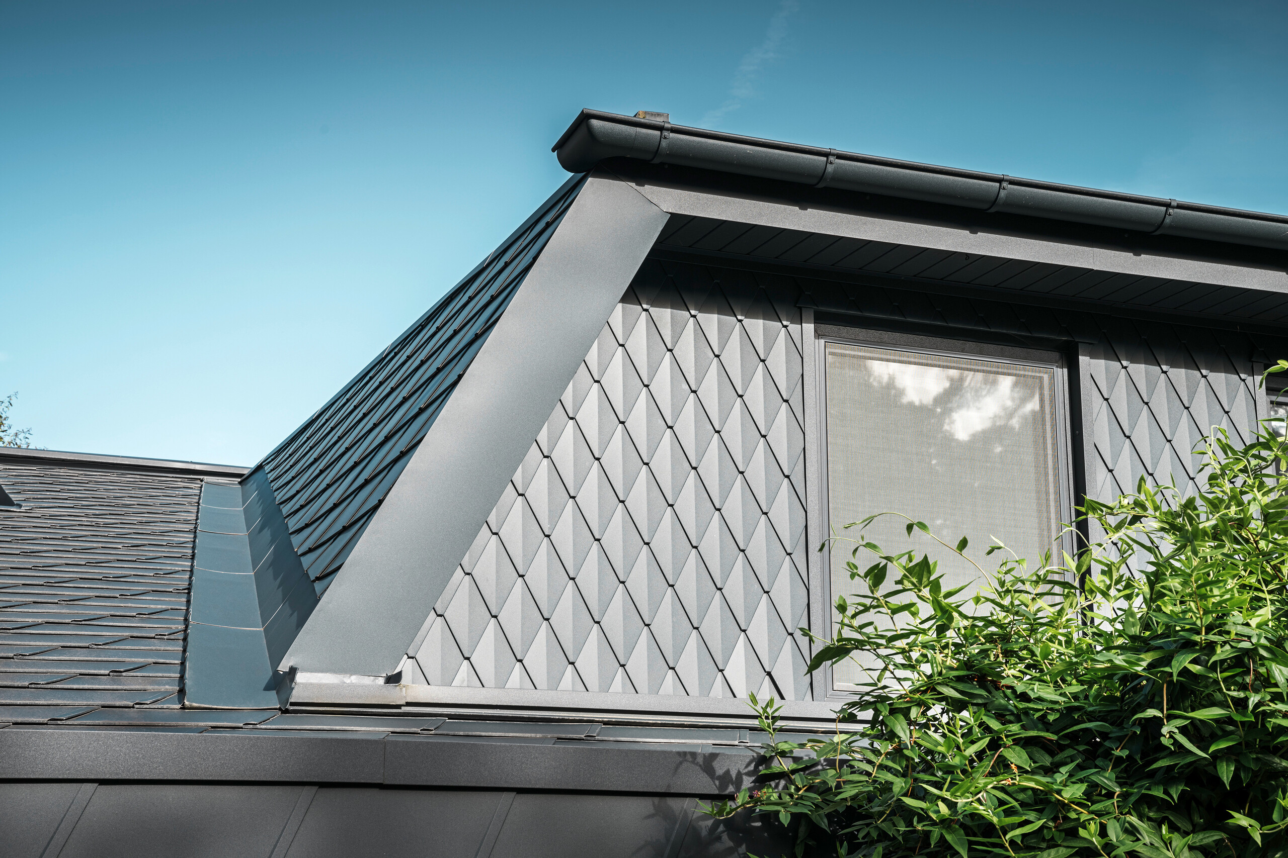 Trapézová vikýřová střecha rodinného domu v Mechernichu, obložená malými falcovanými šablonami PREFA v barvě P.10 antracitová. Střecha je pokryta falcovanými šindeli PREFA DS.19 ve stejné barvě. Vysoce kvalitní hliníkové obložení zajišťuje odolnost proti povětrnostním vlivům a moderní, estetický vzhled.