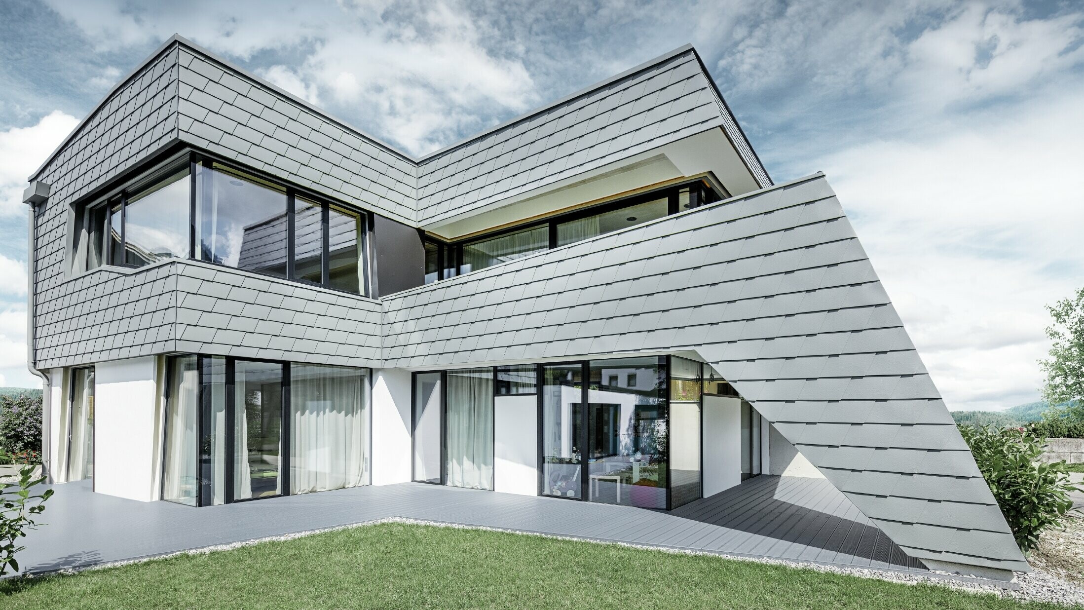 Moderní rodinný dům s plochou střechou, velkými okny a fasádou z hliníkových šindelů v barvě světle šedá od PREFA.
