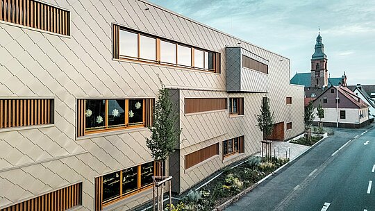 Boční pohled na školku v Alzenau představuje výrazný kontrast k historické městské krajině s pozdně gotickým hradním kostelem v pozadí. Fasáda, obložená bronzovými stěnovými panely PREFA (44 x 44 cm), vytváří elegantní, rytmický vzor a sebevědomě vyniká z okolí – aniž by byla rušivá. Velkorysné okenní otvory s vertikálními dřevěnými žaluziemi a strukturovaná zeleň podél budovy podtrhují koncept udržitelného designu. Kombinace inovativní hliníkové fasády, ekologické konstrukce a citlivosti urbanistického plánování činí z projektu ukázkový příklad zodpovědné architektury, která propojuje tradici a budoucnost.