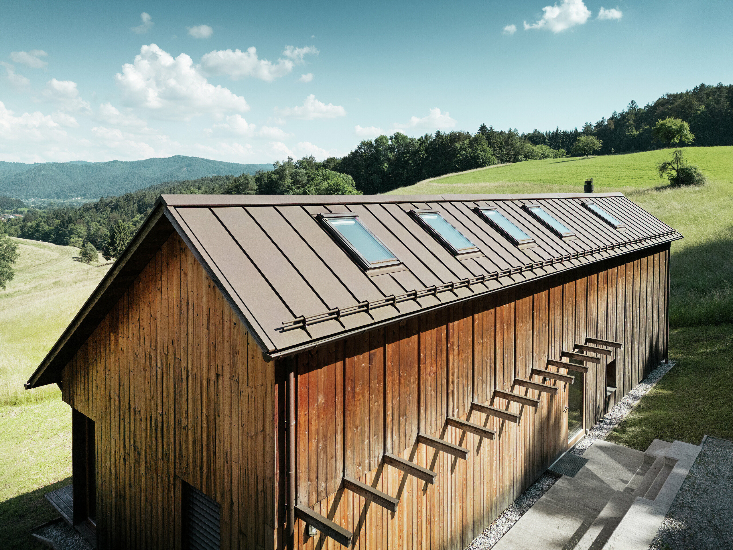 Architektonicky čistý dřevěný dům s vysoce kvalitní hliníkovou střechou PREFALZ v tmavě hnědé barvě P.10. Nenáročná střecha PREFA zaujme svými přesnými liniemi, vysokou odolností proti UV záření a korozi a perfektně integrovanými střešními okny. Kombinace udržitelné dřevěné fasády a odolné falcované střechy harmonicky zapadá do idylické kopcovité krajiny. Asymetrická sedlová střecha má pět přesně umístěných střešních oken, která zajišťují světlo do interiéru.