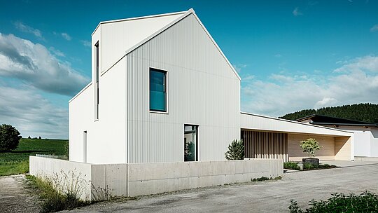 Tento moderní samostatně stojící dům ve Freistadtu je prezentován z dynamické perspektivy s důrazem na čisté linie a výraznou architekturu. Čistě bílý hliníkový obklad PREFALZ se plynule táhne přes celou fasádu a střechu a dodává domu moderní a nadčasový vzhled. Mezi charakteristické designové prvky patří strmá šikmá střecha, velká okna a výrazné vikýře, zatímco svislé drážky na fasádě dodávají domu zajímavou texturu a hloubku. Kontrast s okolní přírodou spolu s elegantními dřevěnými prvky u vchodu a garážových vrat dotváří design. PREFALZ poskytuje obzvláště odolný, nenáročný na údržbu a odolný povrch vůči povětrnostním vlivům, díky čemuž je dům nejen esteticky příjemný, ale také funkční a udržitelný.