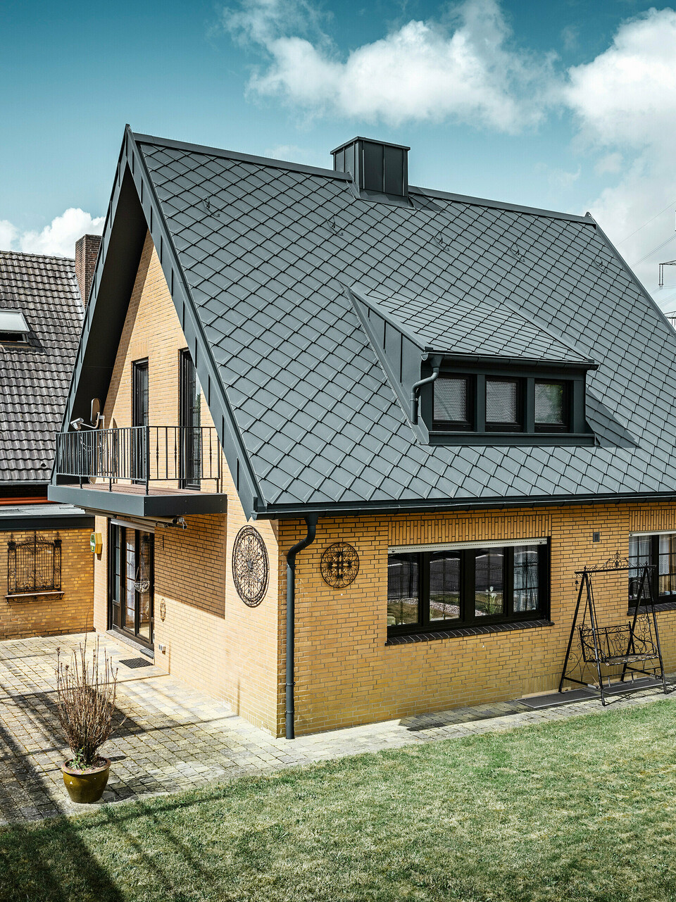 Ein Einfamilienhaus in Tönisvorst mit einem PREFA Dach aus Dachrauten 29×29 in P.10 Anthrazit, das ein herausstechendes Rautenmuster bietet. Schleppgauben, Kamin und der Eingangsbereich sind mit PREFALZ verkleidet, was zusammen mit Ablaufrohr und Hängerinne in P.10 Anthrazit dem Haus einen zeitlosen und eleganten Look verleiht. Das Backsteingebäude mit dem Aluminiumdach harmoniert mit der ruhigen, grünen Umgebung der Siedlung.
