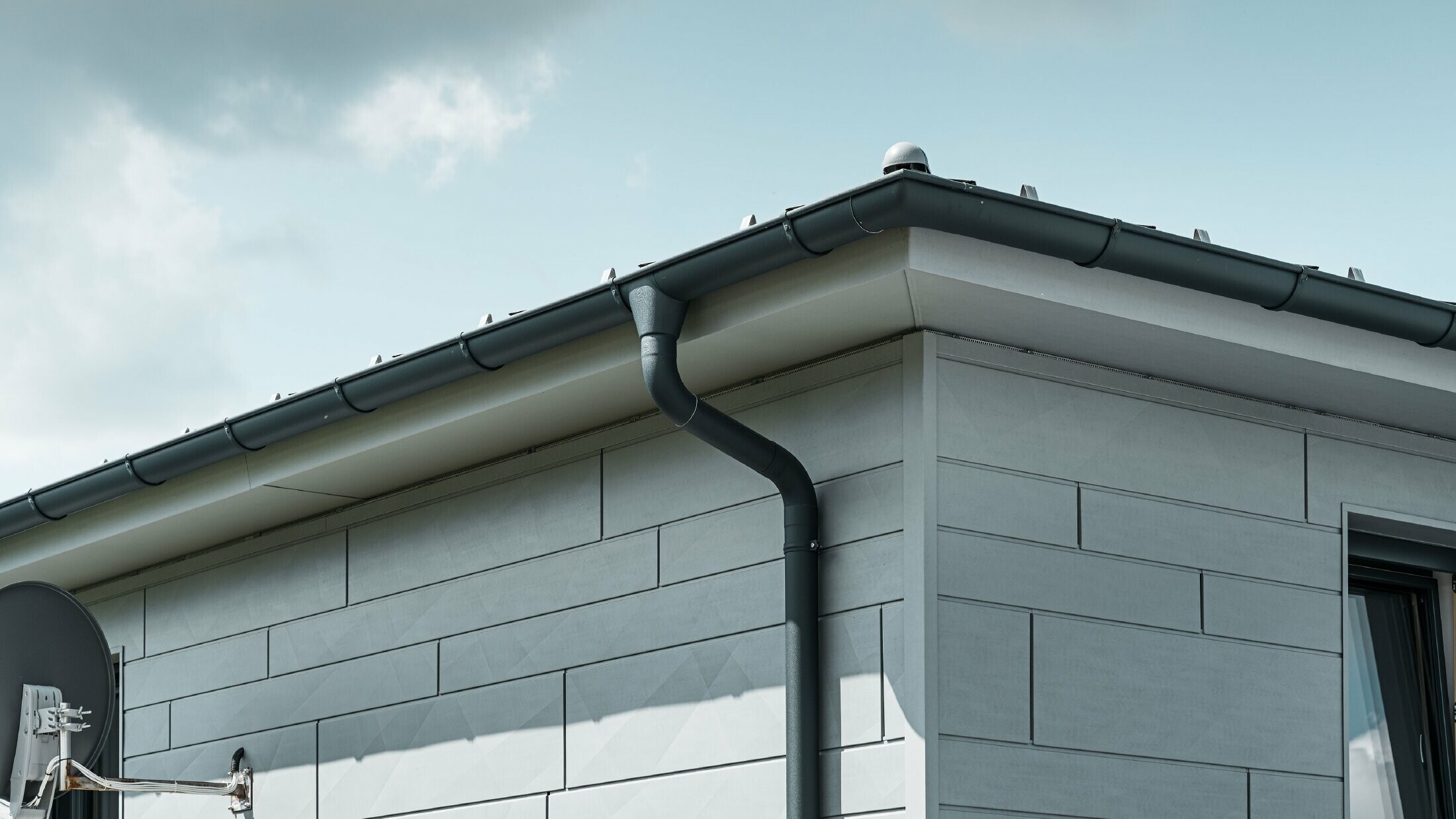 Pohled na střechu domu se střešním žlabem PREFA, kulatým kotlíkem a kruhovým svodem v antracitové barvě a s fasádou z lamel PREFA Siding.X v barvě patina šedá