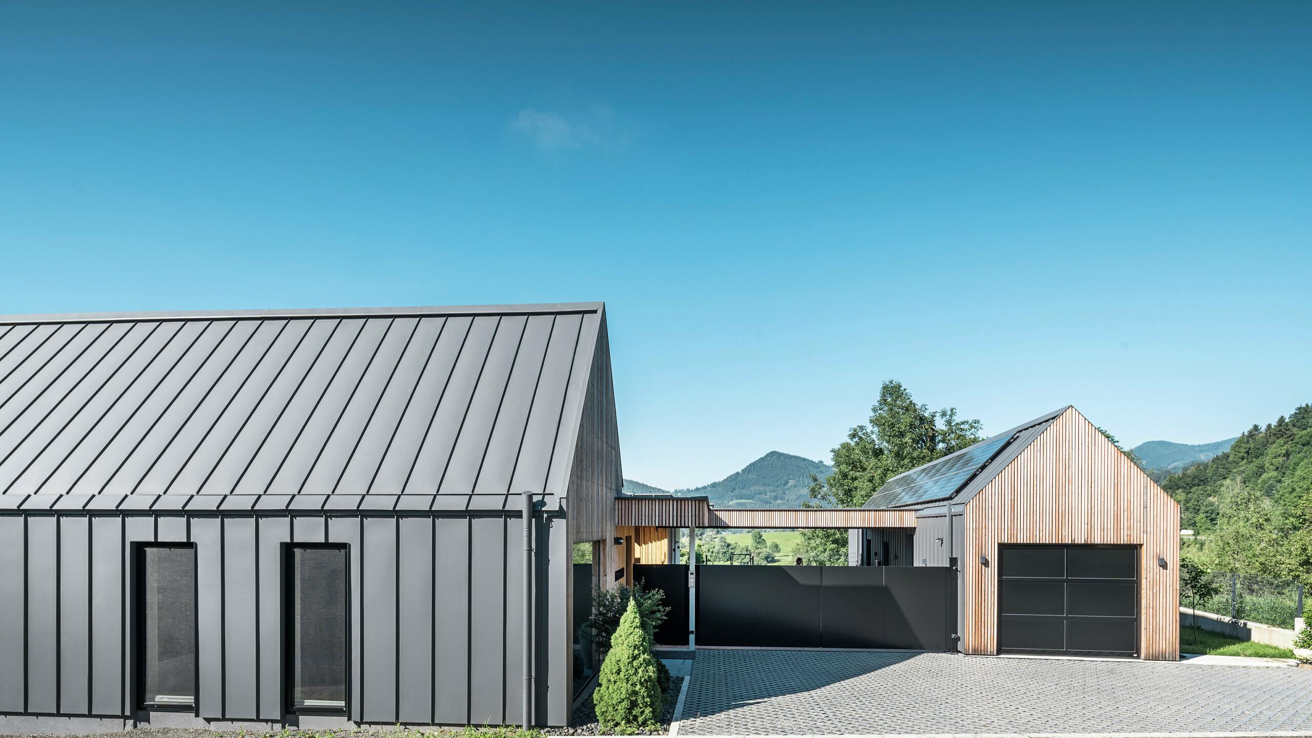 Vstupní areál rodinného domu s přilehlou garáží v rakouském Pogier uprostřed malebné krajiny. Střecha a fasáda obou budov jsou vybaveny odolným PREFALZ v P.10 tmavě šedé barvě, který vytváří moderní vzhled. Garáž se vyznačuje harmonicky integrovaným dřevěným obkladem, který hladce zapadá do okolní přírody. Dlážděné nádvoří a čisté linie designu doplňují estetiku nemovitosti, zatímco v pozadí zvlněné kopce dotvářejí obraz klidné zelené oázy v zeleném srdci Rakouska.