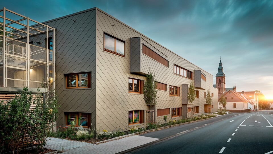 Zalitá měkkým světlem západu slunce, odhaluje školka v Alzenau svou nejpůsobivější stránku. Fasáda, obložená bronzovými stěnovými panely PREFA (44 x 44 cm), odráží večerní světlo a zdůrazňuje vysoce kvalitní, texturovaný povrch moderní hybridní budovy. Jasně strukturovaná architektura se stupňovitými okenními pásy a svislými dřevěnými žaluziemi vytváří výrazný kontrast s historickým kostelem v pozadí. Vlevo dodává jemná kovová konstrukce vnějšímu schodišti nádech transparentnosti. Dobře udržovaný zelený pás podél budovy podtrhuje její závazek k udržitelnosti. Tento obraz působivě kombinuje moderní design budovy, udržitelnou hliníkovou technologii PREFA a harmonické začlenění do historické městské krajiny Alzenau.