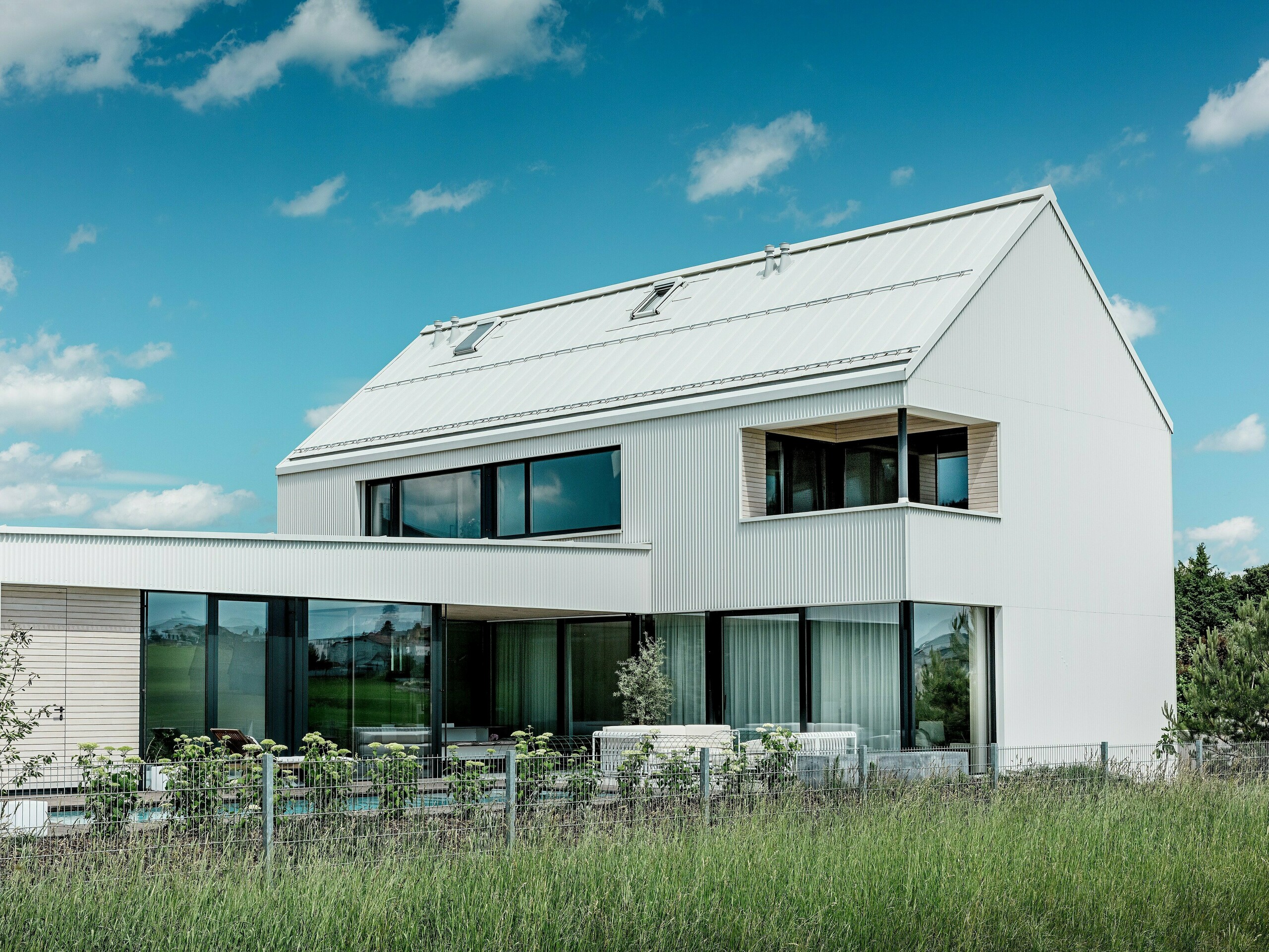 Tento impozantní samostatně stojící dům ve Freistadtu zaujme svou elegantní architekturou s čistě bílým hliníkem PREFALZ na fasádě a střeše. Klikatě skládaná fasáda vytváří moderní, geometrický vzhled, který harmonicky zapadá do okolní přírody. Velká okna nejenže přinášejí do interiéru dostatek přirozeného světla, ale také nabízejí rozlehlý výhled na okolní krajinu. Strmá šikmá střecha a moderní balkon s dřevěným obložením podtrhují minimalistický design a udržitelnou konstrukci domu. PREFALZ zajišťuje vizuální trvanlivost a odolnost vůči povětrnostním vlivům, čímž zaručuje dlouhou životnost s minimální údržbou.