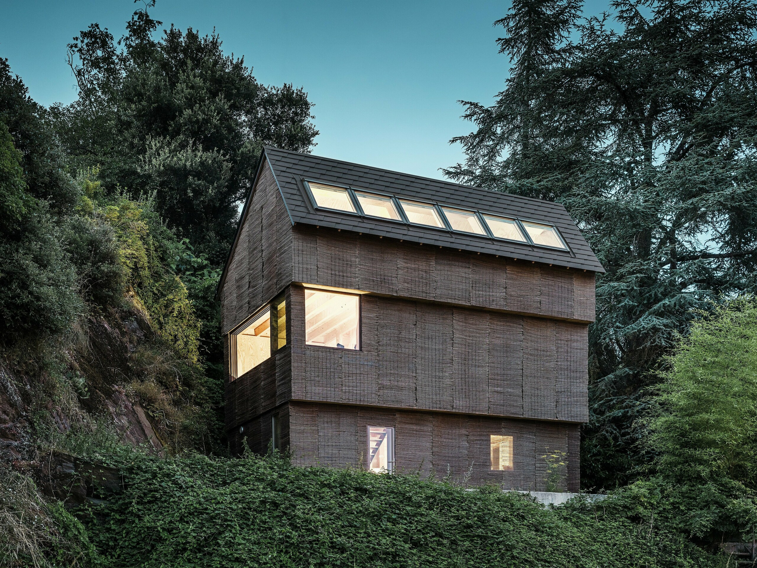 Noční pohled na moderní dům v západní Francii s falcovanými šindeli PREFA v barvě P.10 oříškové barvě a velkorysými střešními okny a udržitelnou proutěnou fasádou