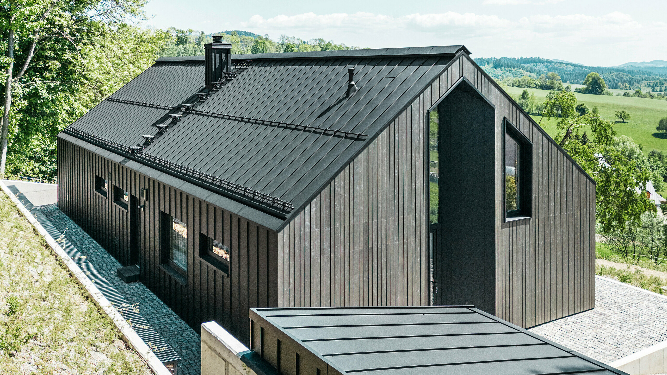 Tmavý dům z masivního dřeva s PREFALZ v P. 10 na střeše a zadní straně budovy. Před domem je malá přístavba s hliníkovým pláštěm z PREFALZ v barvě P.10 černé. V pozadí je jasně modrá obloha a les.