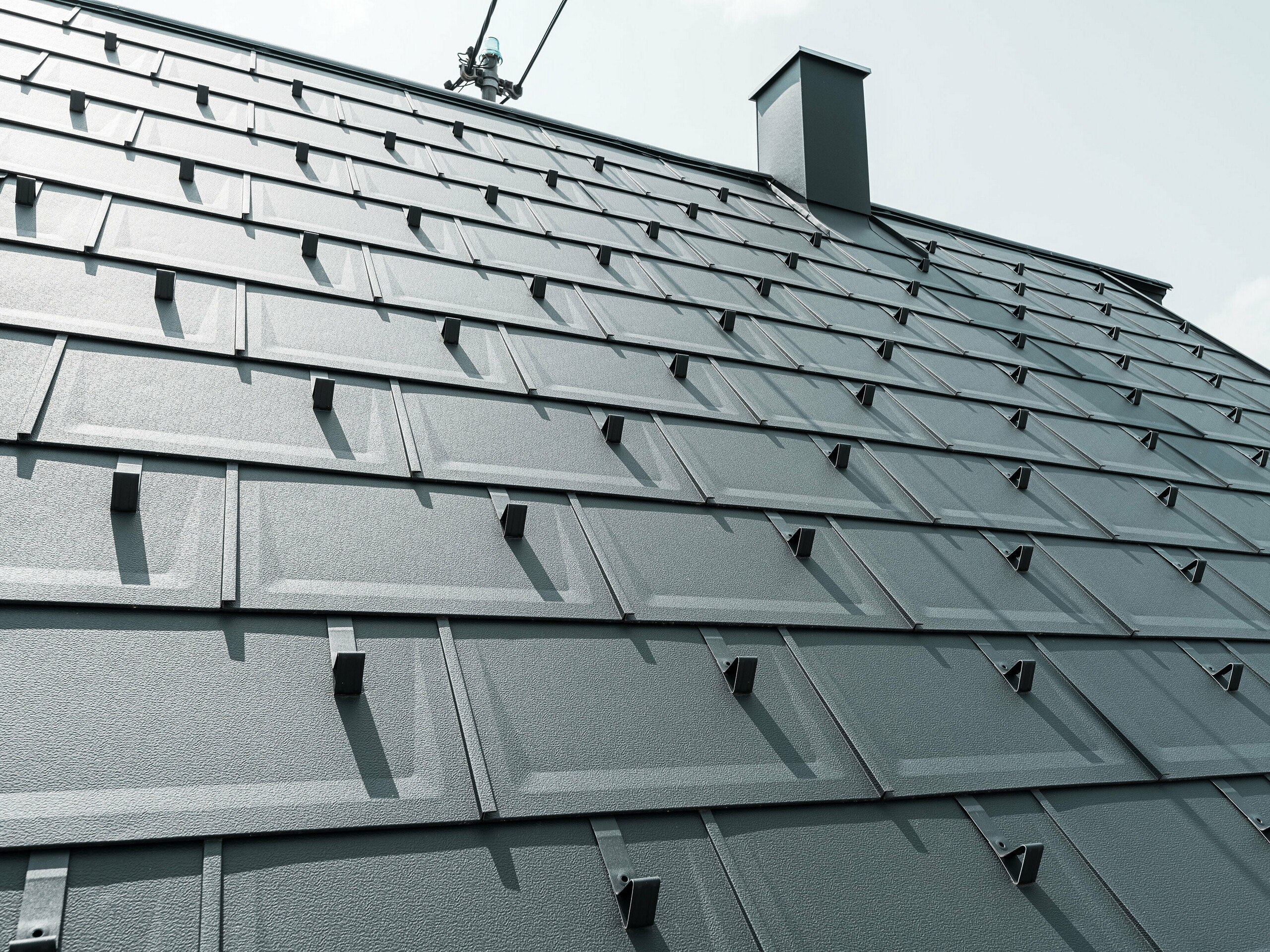 Detailní záběr střešní panelu PREFA R.16 v barvě P.10 antracitová. Na střeše jsou vidět sněhové zábrany PREFA a komínová krytina.