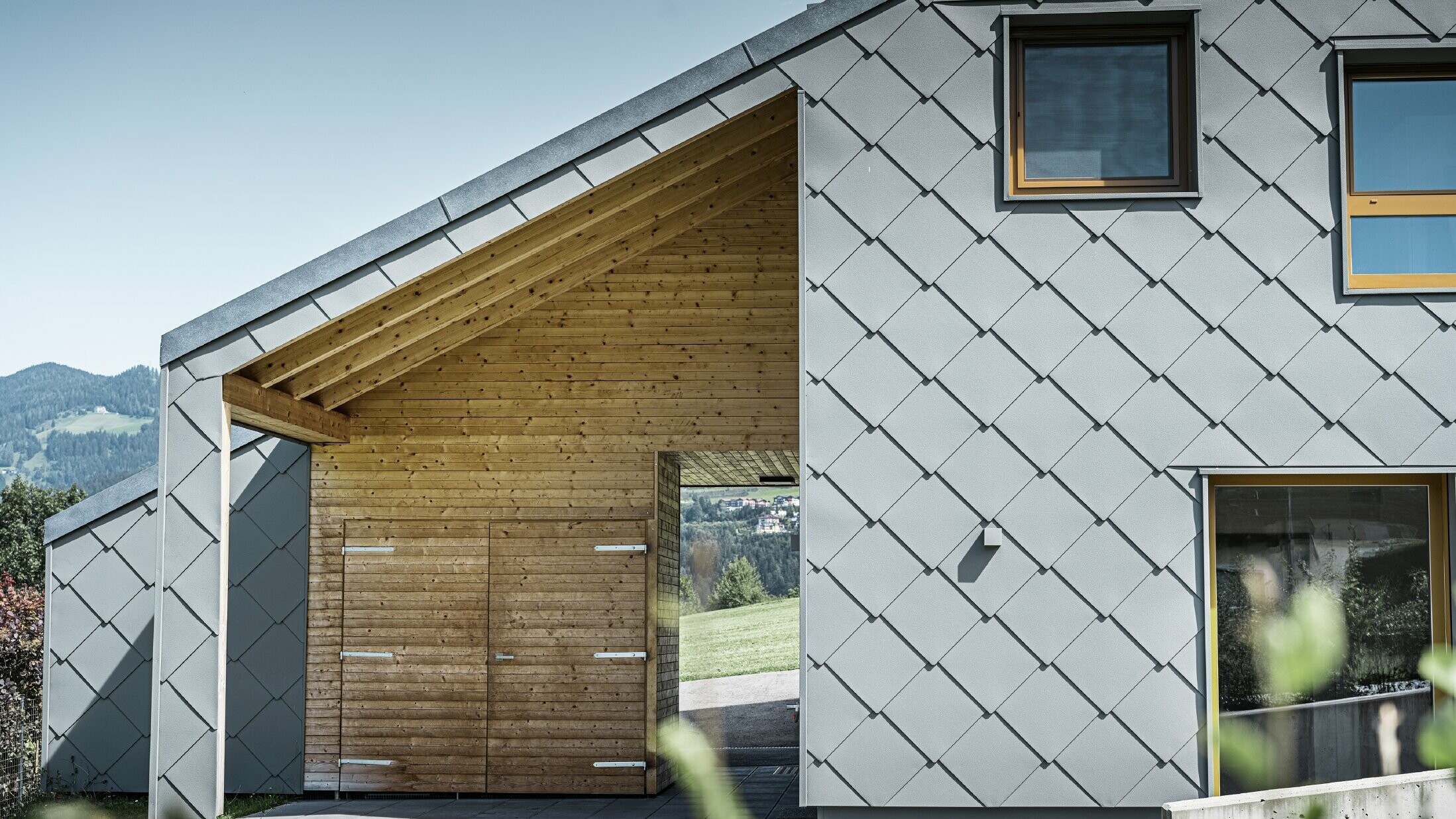 Krytá vstupní část je obložená vodorovným dřevěným obkladem, zbytek fasády je obložen velkými PREFA fasádními šablonami z hliníku ve světle šedé barvě