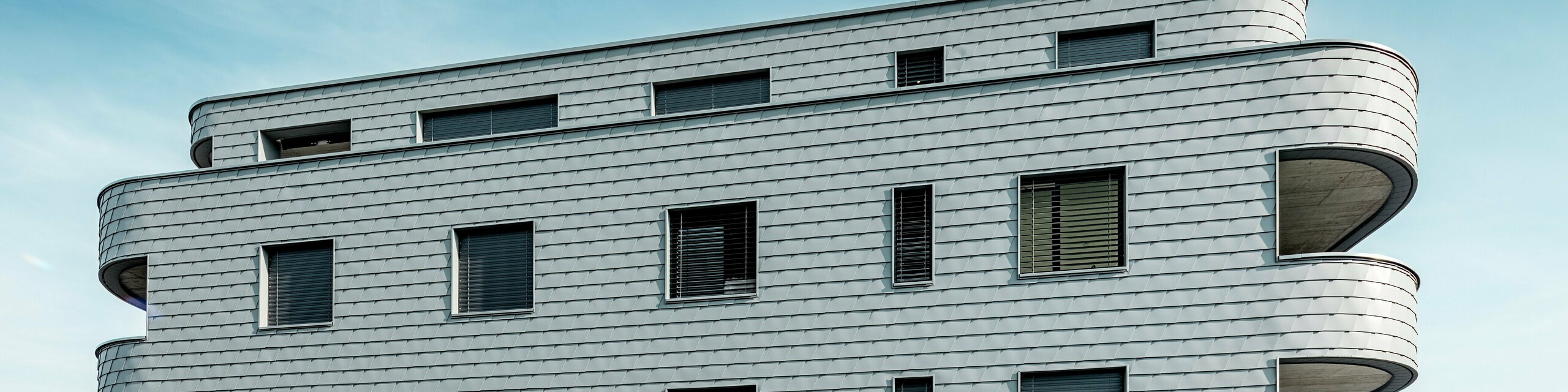 Čelní pohled na Bernerhaus v Buchsu s jemně zaoblenými rohy budovy a souvislou fasádou z PREFA fasádních šindelů v barvě P.10 světle šedé. Šupinatá hliníková fasáda vytváří zářivý vzor a zdůrazňuje horizontální linie zakřivených balkonových pásů. Precizně integrované okenní otvory harmonicky splývají s homogenním povrchem. Díky lehkému, povětrnostním vlivům odolnému a trvanlivému hliníku PREFA vzniká nenáročný, rozměrově stabilní plášť budovy s výrazným vzhledem na alpském pozadí.