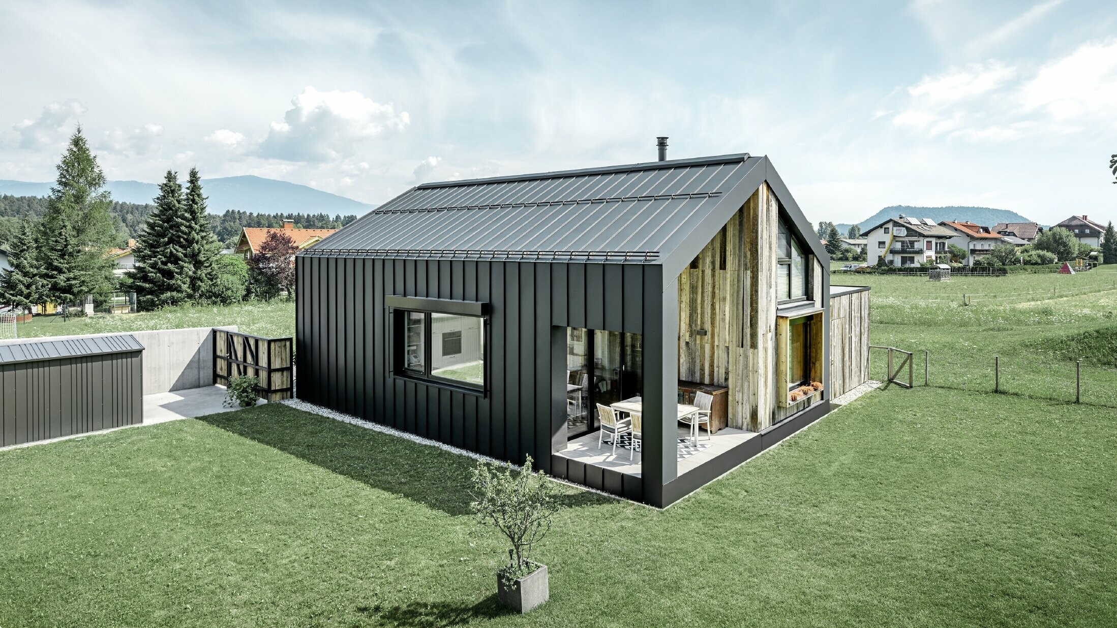 Rodinný dům se sedlovou střechou, střecha a fasáda jsou pokryty PREFALZem společnosti PREFA v provedení P.10 antracitová. Šáry plochy střechy přecházejí ve fasádu. Část fasády je obložena zvětralým dřevem.