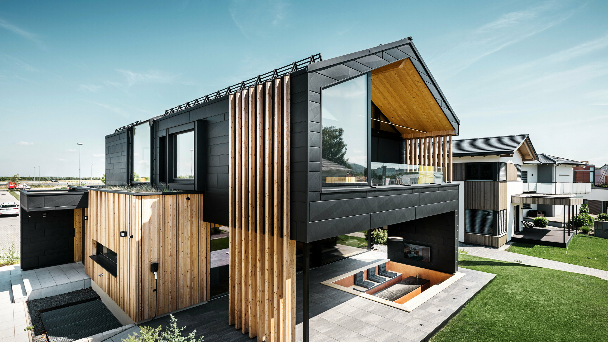 [Translate to Český:] Seitenansicht des Musterhauses Autark in der Blauen Lagune in Vösendorf, Österreich. Das Obergeschoss besticht durch große Fensterflächen und eine moderne Aluminiumfassade aus PREFA Siding.X Paneelen in P.10 Schwarz, kombiniert mit vertikalen Holzelementen. Die großzügige Terrasse im Erdgeschoss mit eingelassener Sitzgelegenheit fügt sich nahtlos in die Architektur ein. Das stilvolle und nachhaltige Design des Hauses verbindet Aluminium und Holz auf eindrucksvolle Art und Weise.