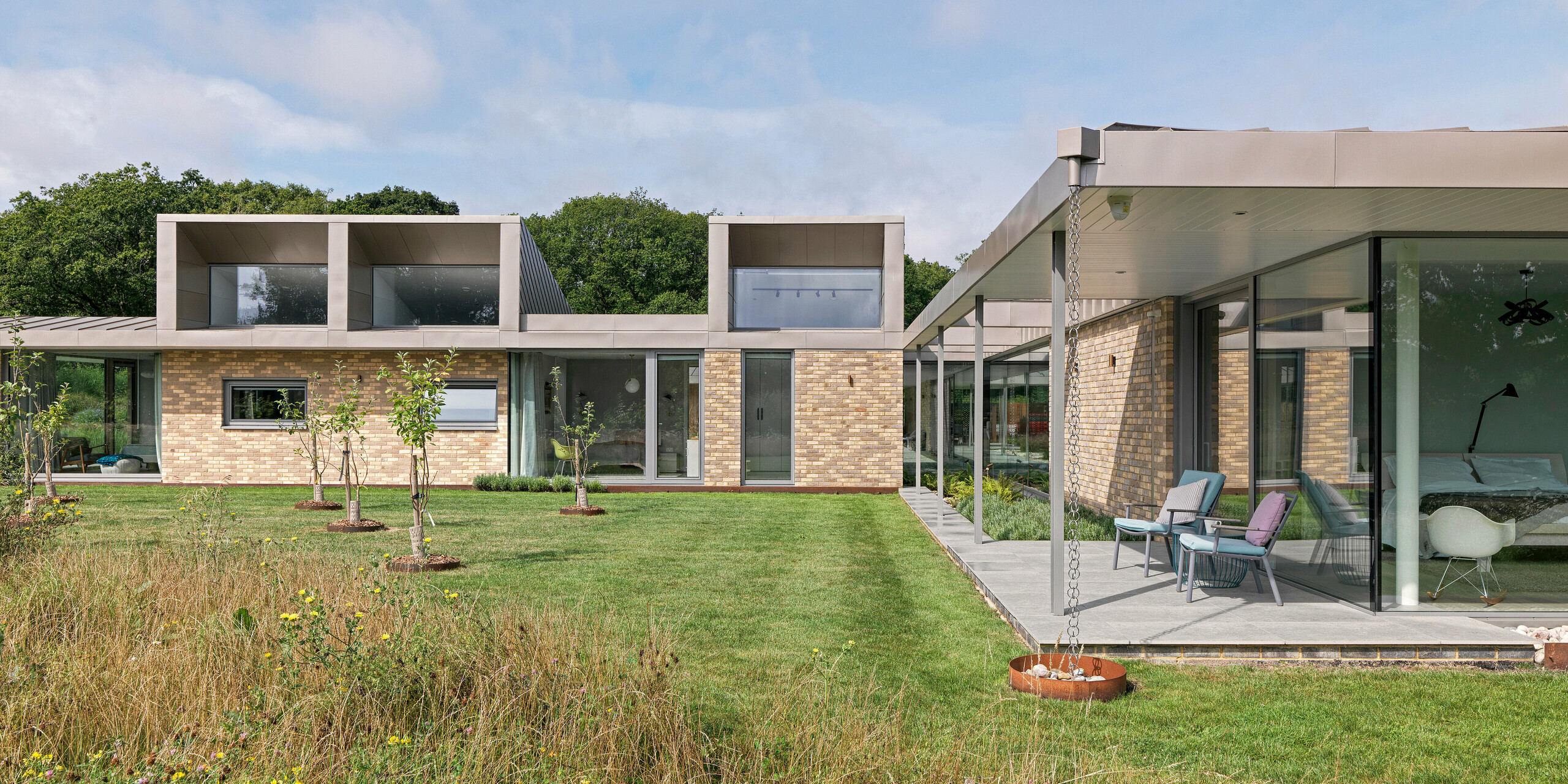 Seitenansicht eines luxuriösen Wohnhauses in West Sussex mit markanten Aufbauten im Obergeschoss, verkleidet mit PREFA Wandrauten 20 × 20 in P.10 Bronze. Das Flachdach besteht aus PREFALZ Aluminium, ebenfalls in P.10 Bronze, und unterstreicht das minimalistische Design. Großflächige Glasfassaden, eine überdachte Terrasse mit filigranen Stützen sowie Regenablaufketten runden das nachhaltige Architekturkonzept mit langlebigen und wartungsfreien PREFA Aluminiumprodukten ab.