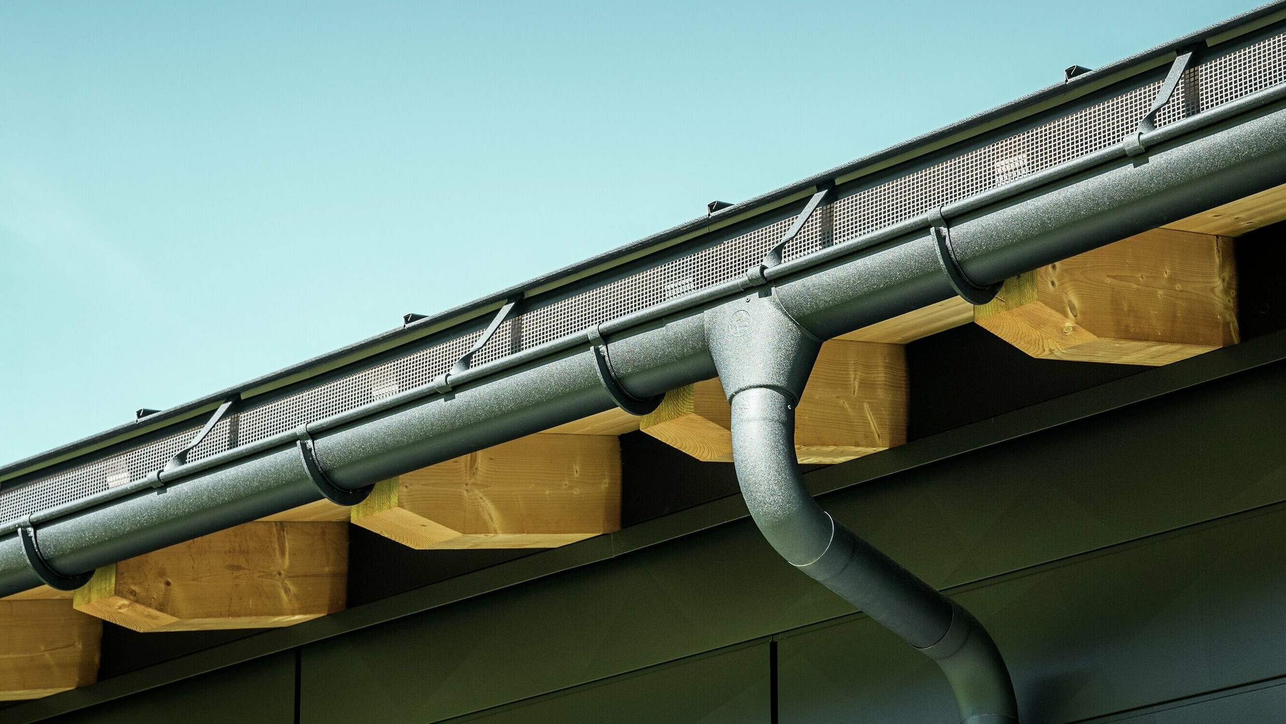 Detailní záběr fasády moderního rodinného domu v Ascharině ve Švýcarsku. Stěny budovy byly obloženy výraznými fasádními panely PREFA Siding.X v barvě P.10 černá. Barevně sladěný je také okapový systém PREFA s půlkulatým žlabem a kruhovým svodem.