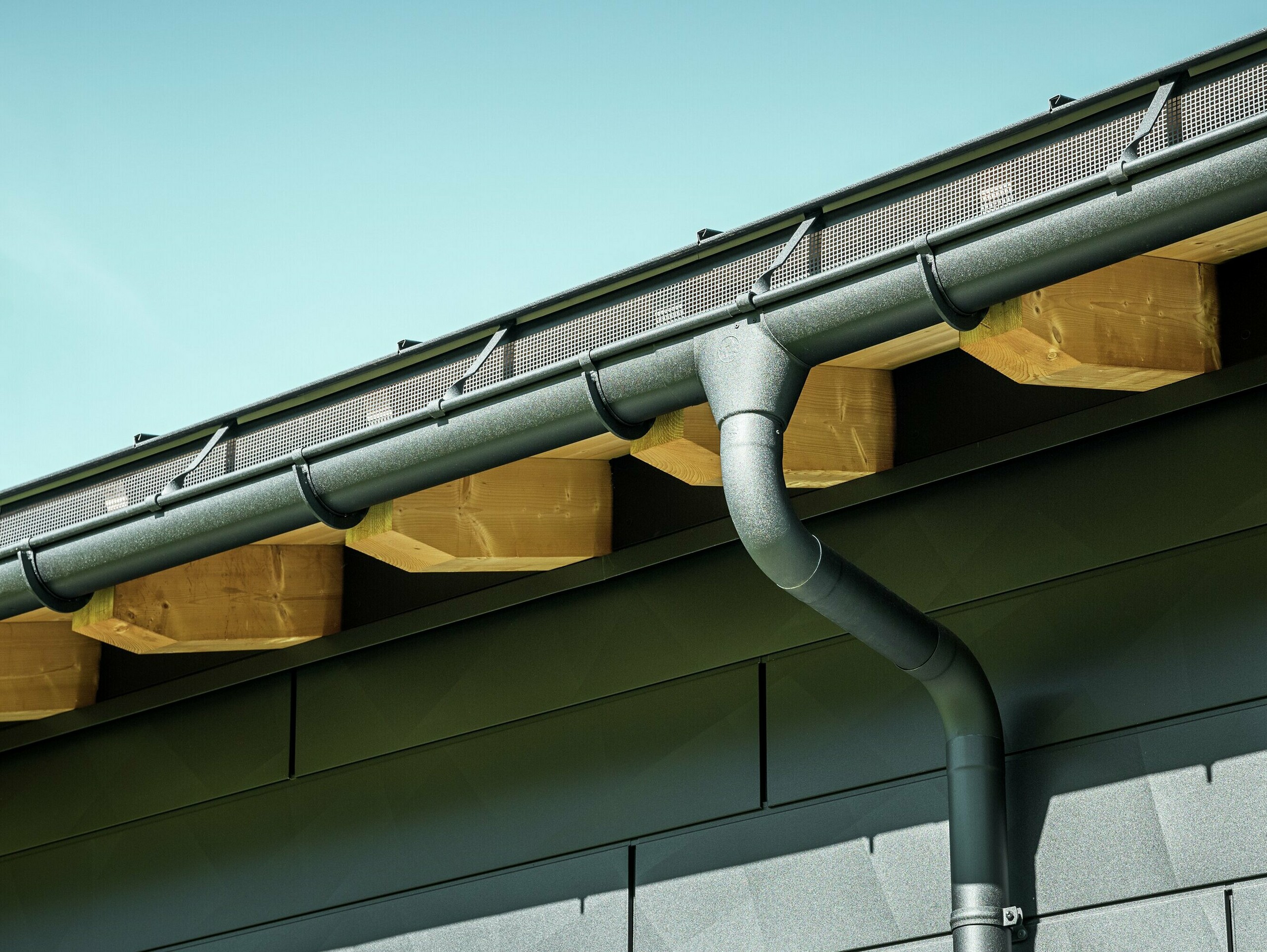 Detailní záběr fasády moderního rodinného domu v Ascharině ve Švýcarsku. Stěny budovy byly obloženy výraznými fasádními panely PREFA Siding.X v barvě P.10 černá. Barevně sladěný je také okapový systém PREFA s půlkulatým žlabem a kruhovým svodem.