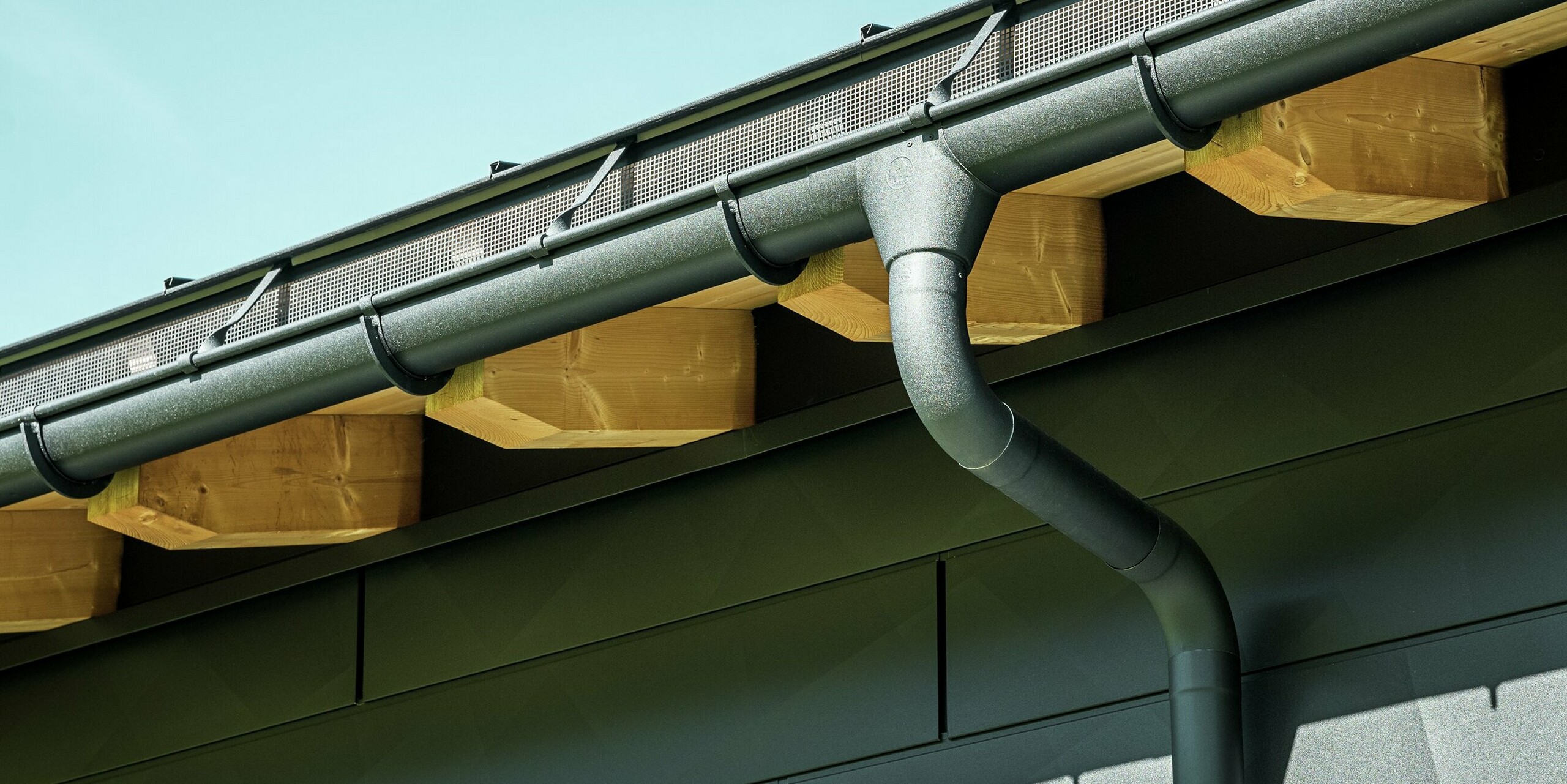 Detailní záběr fasády moderního rodinného domu v Ascharině ve Švýcarsku. Stěny budovy byly obloženy výraznými fasádními panely PREFA Siding.X v barvě P.10 černá. Barevně sladěný je také okapový systém PREFA s půlkulatým žlabem a kruhovým svodem.