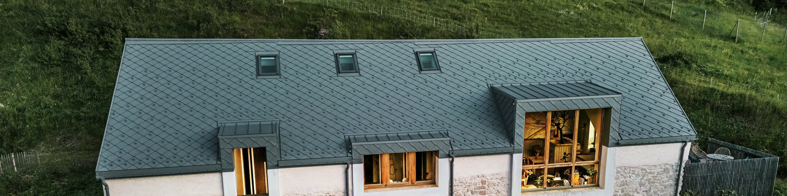 Večerní pohled na zrekonstruovanou stodolu "La Grange à Gaby" v Engins, Francie. Budova kombinuje tradiční kamenné zdivo s moderní hliníkovou střechou PREFA z falcovaných šablon 29 × 29 cm. Velká okna a prosvětlené interiéry podtrhují přívětivou atmosféru nového kulturního centra.