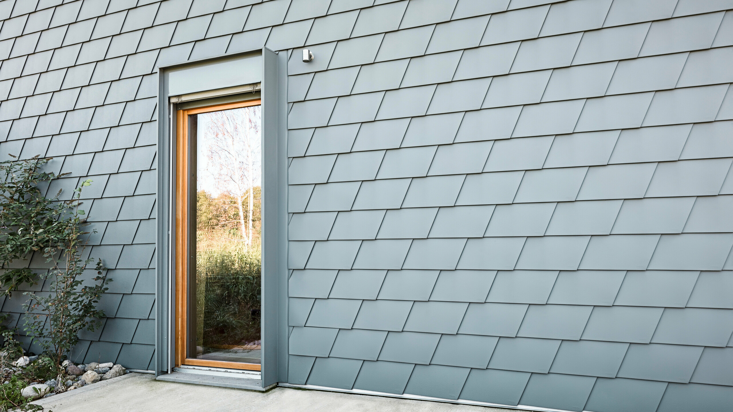 Pohled na fasádu Casa Longaretti s falcovanými šindeli PREFA v barvě P.10 světle šedá, které vytvářejí šupinatou texturu a dodávají budově moderní charakter. Harmonické barevné provedení a přesná pokládka šindelů podtrhují prémiovou kvalitu a inovativní design hliníkových produktů PREFA. Dřevěný rám dveří přidává přirozený kontrast, který zdůrazňuje estetický a udržitelný koncept této budovy.