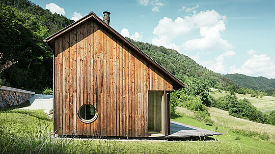 Minimalistický dřevěný dům se zaoblenými okenními otvory a odolnou hliníkovou střechou PREFALZ v barvě P.10 tmavě hnědé zasazený do kopcovité krajiny Vinje. Střešní krytina PREFALZ je bezúdržbová, nabízí optimální ochranu před větrem a povětrnostními vlivy a barevně ladí s přírodní dřevěnou fasádou. Podtrhuje čistou a nadčasovou architekturu budovy. Z prostorné terasy se nabízí úchvatný výhled na kopcovitou slovinskou krajinu. Perspektiva ukazuje přední část rodinného domu s asymetrickou sedlovou střechou a střešními plochami různých velikostí.