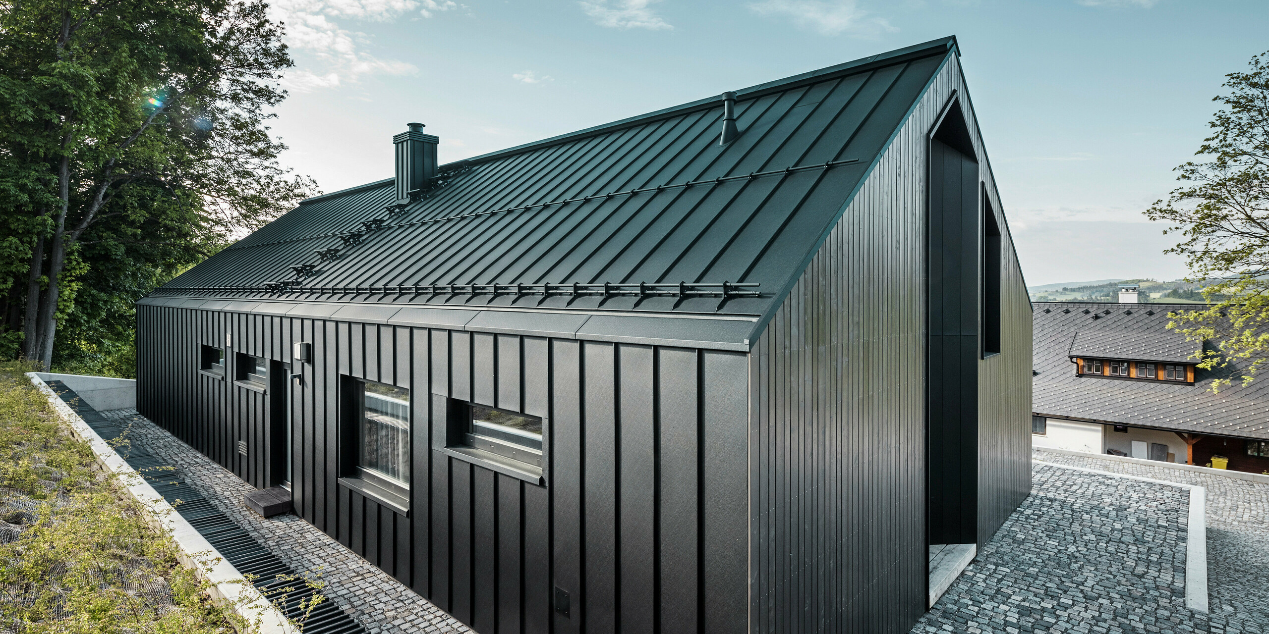 Kombinace černého opláštění domu z hliníku a tmavé dřevěné fasády upoutá pozornost. Díky chytrému začlenění podlouhlého bungalovu do svahu to vypadá, jako by zde stál odjakživa.