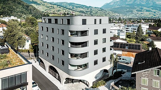 Pohled z ptačí perspektivy na Bernerhaus v Buchsu s jeho výrazným trojúhelníkovým tvarem a jemně zaoblenými balkony zasazenými do alpské krajiny. Souvislá fasáda z PREFA fasádních šindelů ve světle šedé barvě P.10 obaluje budovu jako šupinovitý hliníkový plášť a vytváří živou hru světla a textury. Nad masivním betonovým základem s arkádami působí lehká, povětrnostním vlivům odolná hliníková fasáda PREFA mimořádně precizně, trvanlivě a nenáročně na údržbu – moderní poutač v městské krajině.