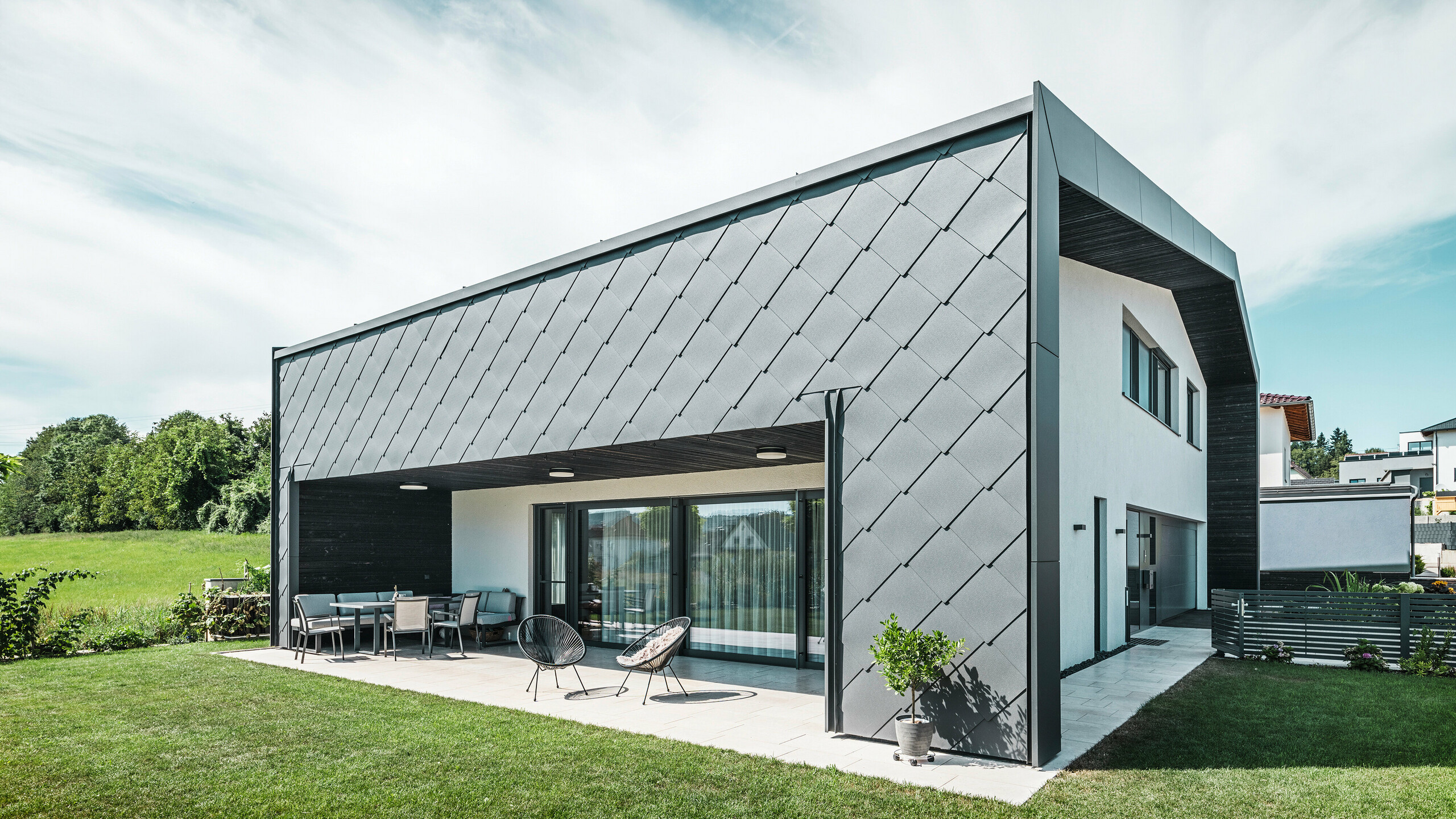 Upravená zahrada rodinného domu v Aschachu. Budova je stylově pokryta tmavě šedými fasádními šablonami PREFA Aluminium 44 × 44. Krytá terasa nabízí příjemné posezení s výhledem na svěží zelený trávník. Elegantní hliníkový obklad v kombinaci s bílými omítkami zajišťuje moderní a povětrnostním vlivům odolnou architekturu. Velké okna propouštějí do interiéru dostatek denního světla a vytvářejí otevřenou, světlou atmosféru.