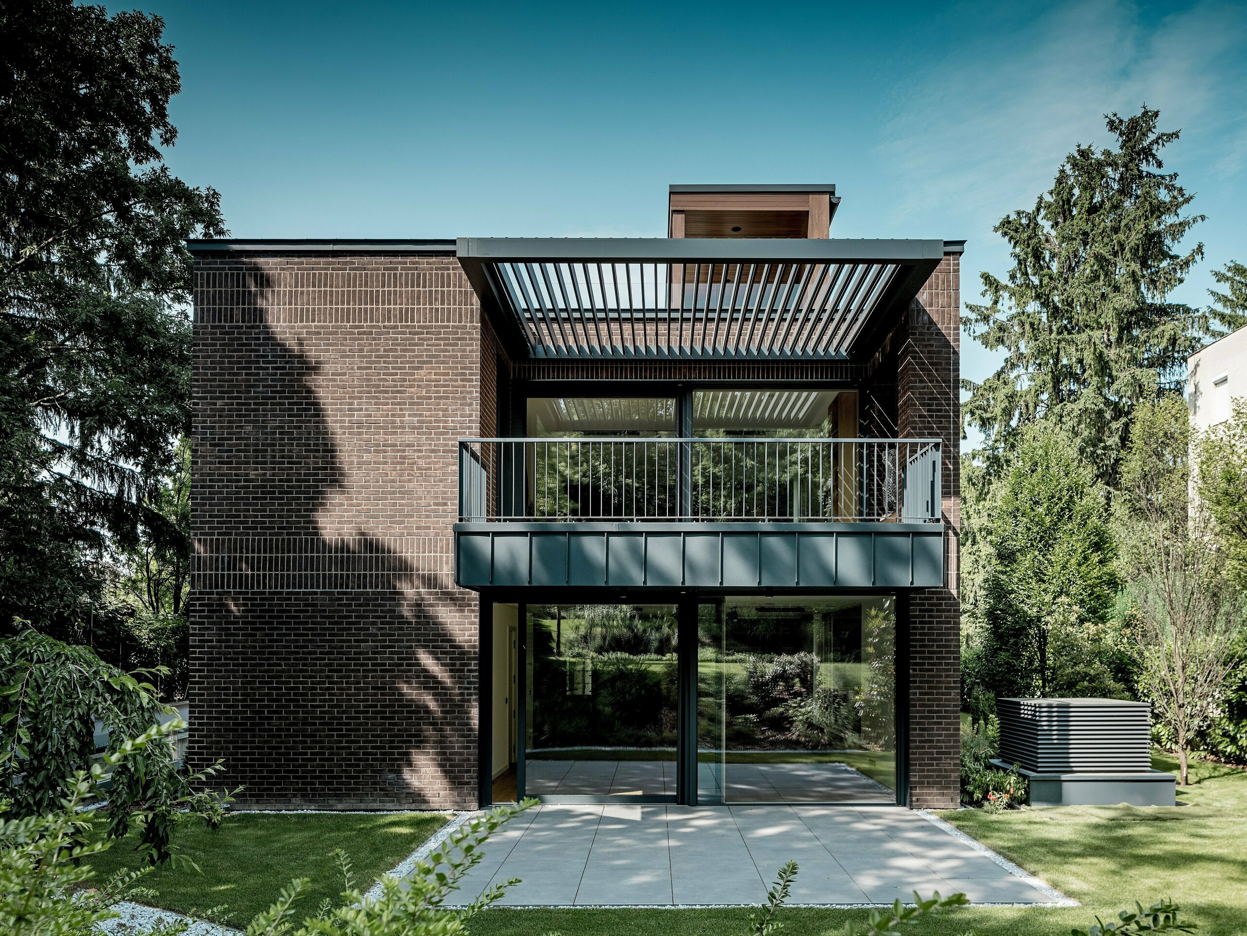 Fotografie ukazuje zahradní stranu moderní, vícegenerační vily v Budapešti, která se vyznačuje jasně strukturovanou fasádou z tmavě červených klinkerových cihel a hliníku PREFALZ v antracitové barvě P.10. Velký balkon s jemnou kovovou konstrukcí a integrovanými žaluziemi vytváří podmanivou hru světla a stínu. Prosklené fasády od podlahy až ke stropu otevírají interiér zelené zahradě. Precizně provedené hliníkové detaily podtrhují trvanlivost, odolnost vůči povětrnostním vlivům a architektonickou jasnost řešení PREFA a dodávají budově výrazný a nadčasový charakter.