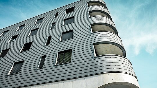 Detailní záběr na Bernerhaus v Buchsu s výrazným zaobleným rohem a plynulými balkonovými otvory. Fasáda, obložená falcovanými šindeli PREFA v barvě P.10 světle šedé, překrývá jasně strukturovaný kubický tvar jako precizně vyrobený hliníkový plášť. Šupinovitý povrch zdůrazňuje měkké křivky a vytváří živou hru světla a stínu. Díky lehkému, povětrnostním vlivům odolnému a trvanlivému hliníku PREFA je dosaženo homogenního, nenáročného pláště budovy – technicky sofistikovaného a architektonicky atraktivního.