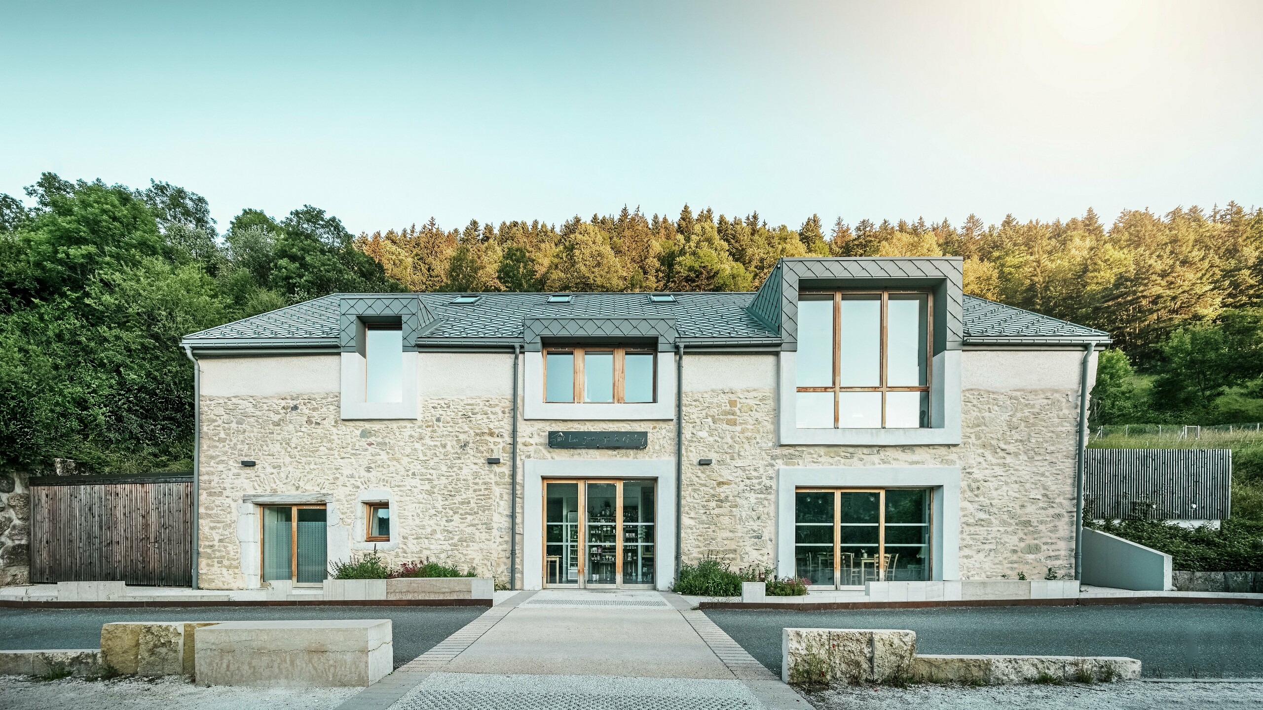 Čelní pohled na obnovenou stodolu "La Grange à Gaby" v Engins, Francie. Kombinace tradičního kamenného zdiva a moderní hliníkové střechy PREFA s falcovanými šablonami 29 × 29 cm dodává budově harmonický a současný vzhled. Stodola dnes slouží jako místo kulturního setkávání a ubytovna.