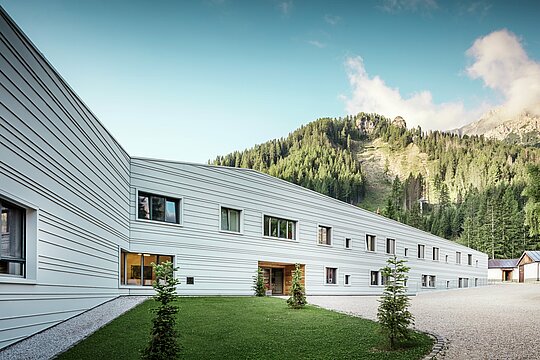 Aluminium trifft auf Präzision – ein harmonischer Dialog zwischen Technologie und Natur. Vor der imposanten Kulisse der Dolomiten präsentiert sich das „Villaggio Alpino Tempesti“ mit klaren Linien und modernem Design. Die PREFALZ-Fassade in der Farbe P.10 Prefaweiß ist nicht nur extrem widerstandsfähig, sondern schafft dank ihrer hellen Farbe durch die Reflexion der Sonnenstrahlen auch ein angenehmes Mikroklima. Grüne Flächen und Holzelemente am Eingang des Militärgeländes erzeugen eine überraschend einladende Atmosphäre.