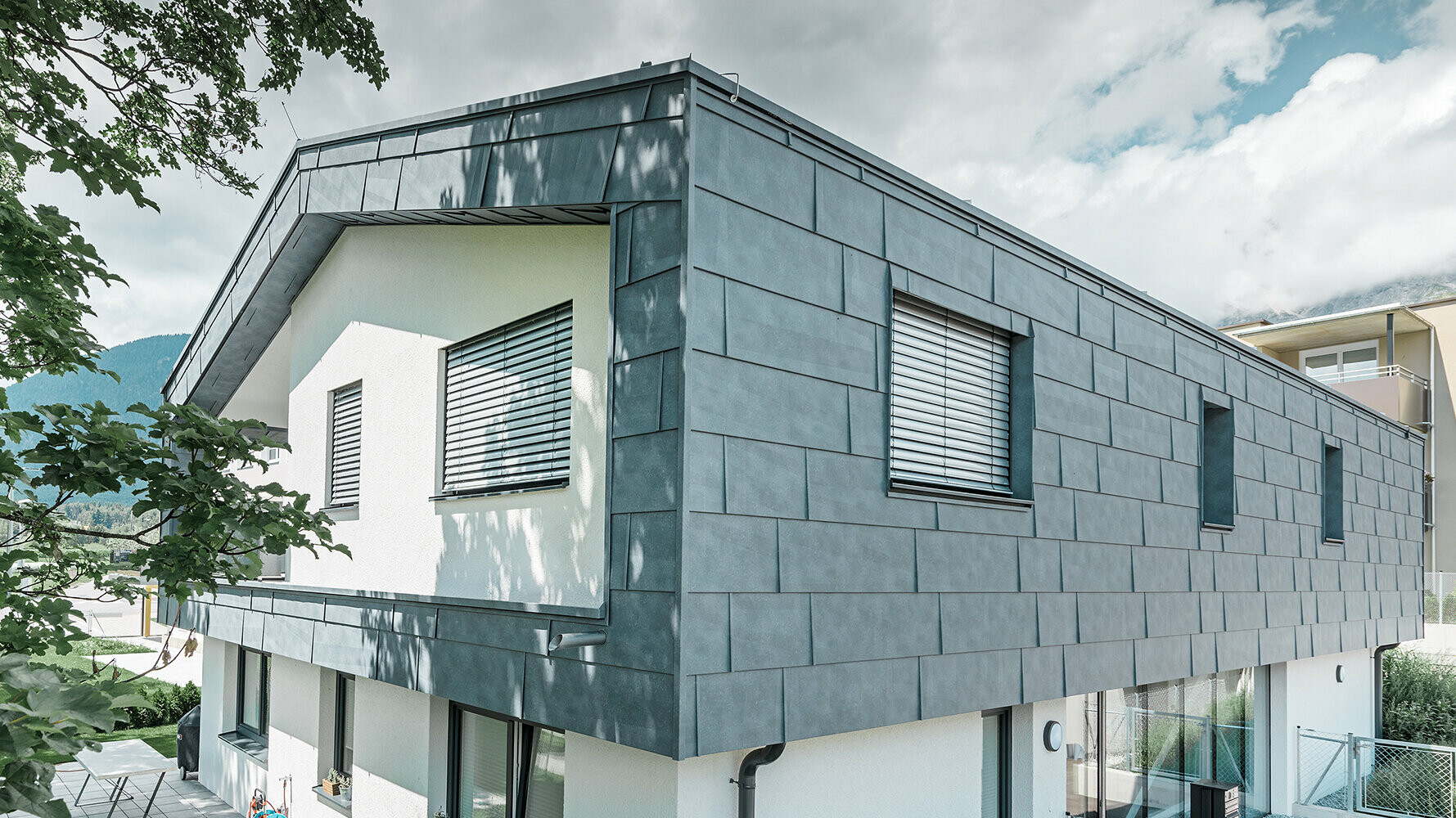 Moderní rodinný dům bez střešního přesahu. Horní poschodí bylo obloženo PREFA fasádními panely FX.12 břidlicové barvy, zbytek fasády má bílou omítku.