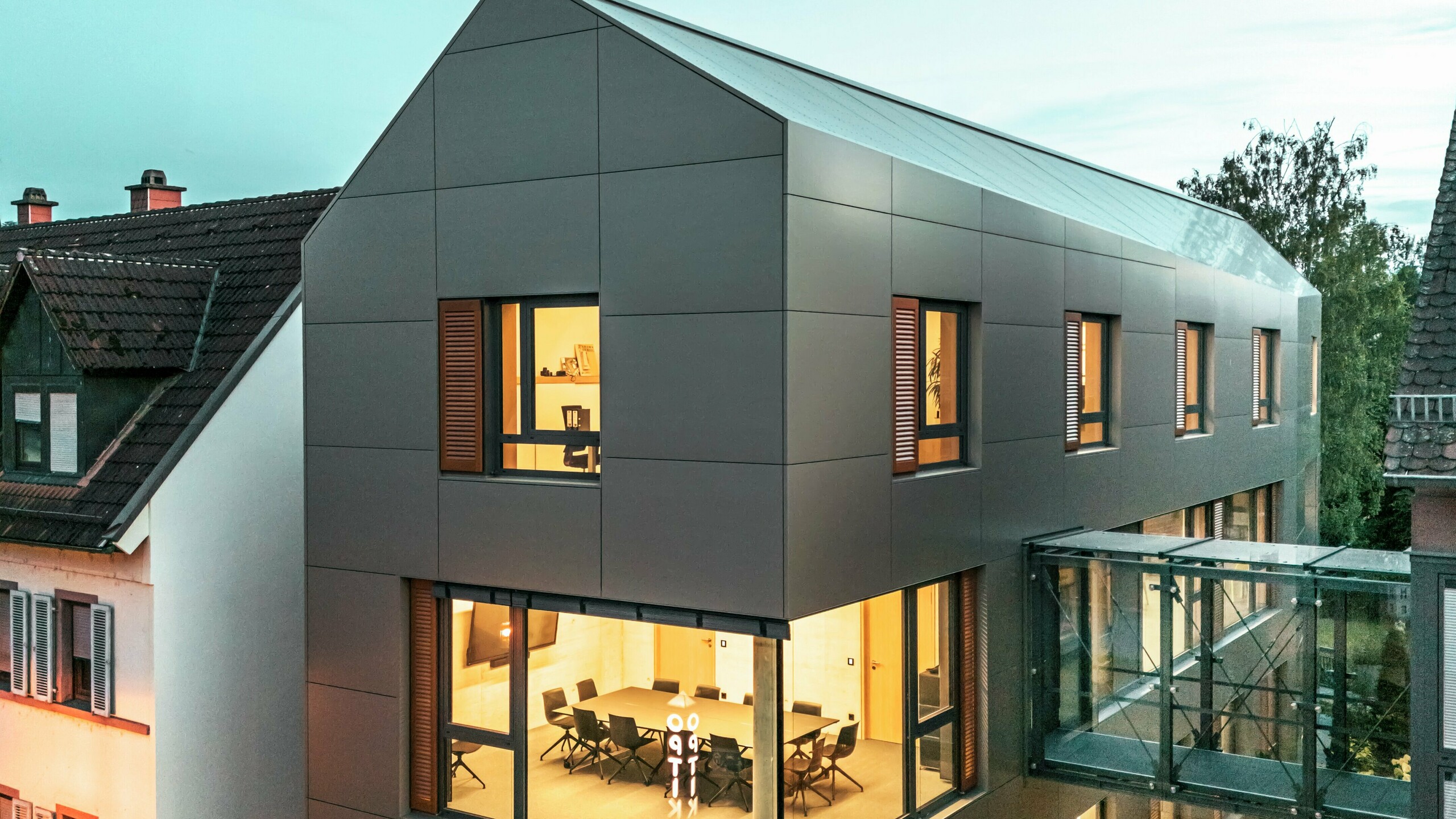 Na obrázku je radnice v Alzenau za soumraku s moderní fasádou z hliníkových kompozitních panelů PREFABOND v elegantním bronzovém odstínu. Skleněná chodba spojující budovu s přilehlou částí vytváří vizuální kontrast. Velká okna umožňují pohled do interiéru, který je osvětlen teplým světlem. Jasný geometrický tvar fasády a okolní zeleň umocňují moderní a současný vzhled budovy.