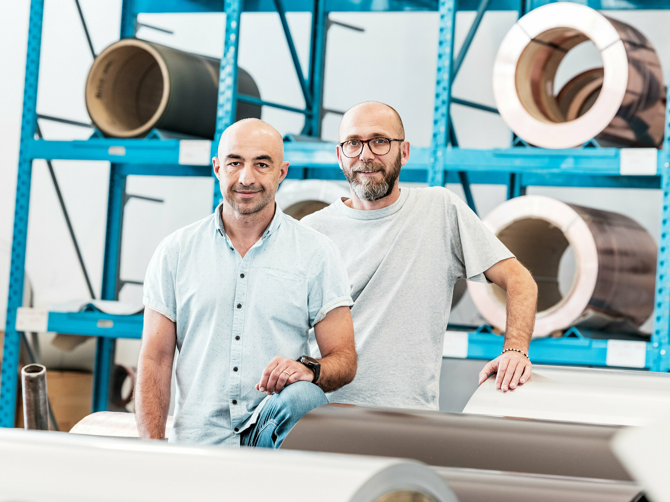 Dva řemeslníci ve skladu před navinutými hliníkovými svitky – moderní pracovní prostředí pro hliníkové střešní a fasádní systémy.