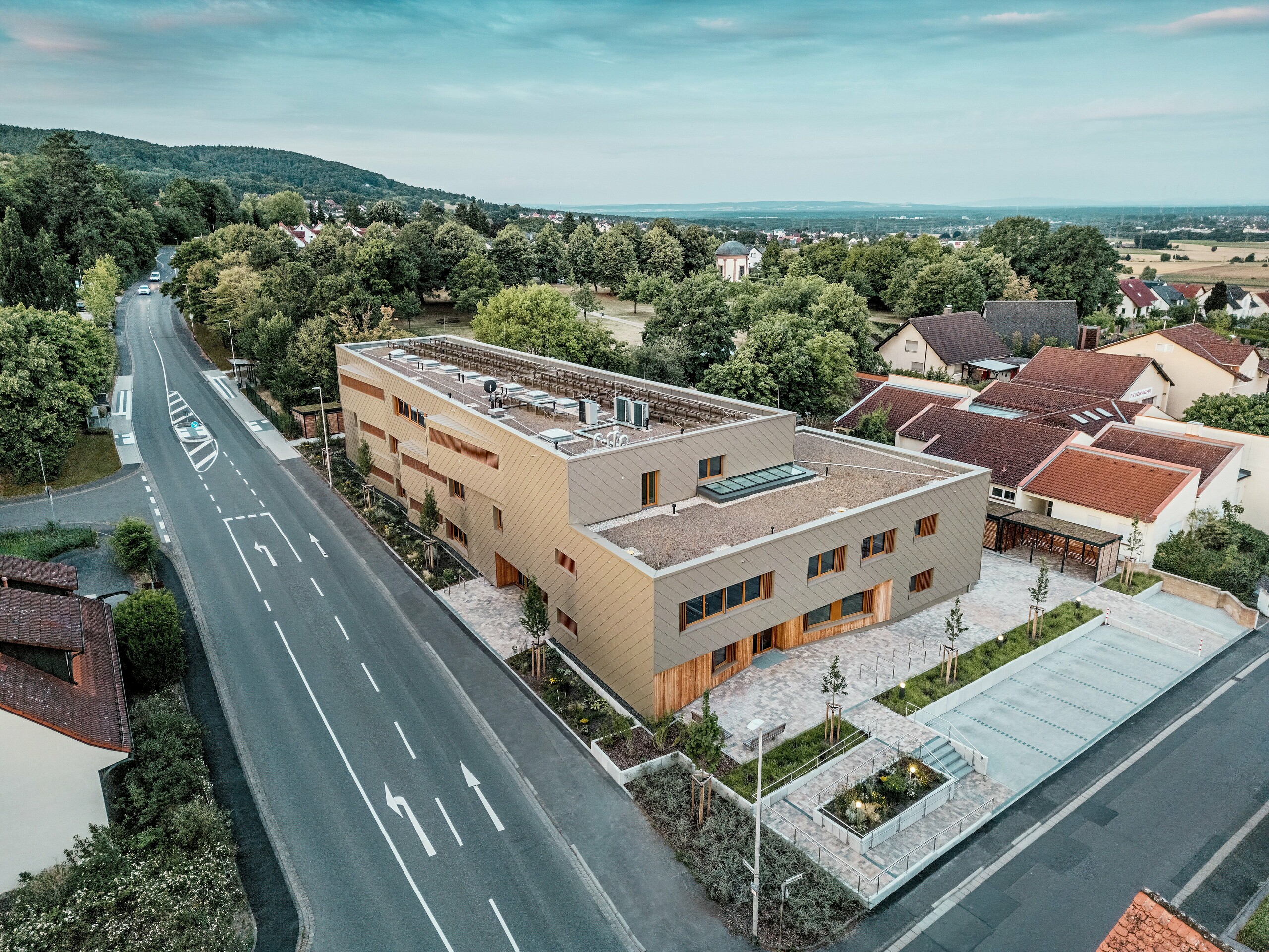 Působivý celkový design městské denní péče v Alzenau je jasně viditelný na leteckém snímku. Hranatá hybridní budova je harmonicky začleněna do klidné obytné čtvrti na okraji lesa Spessart. Fasáda z bronzově zbarvených stěnových panelů PREFA (44 x 44 cm) dodává budově výrazný, vysoce kvalitní vzhled a zdůrazňuje její moderní architekturu. Zelené střechy, prostorné venkovní plochy a dobře navržený dopravní tok podtrhují celkovou udržitelnou koncepci. Kombinace odolného hliníku, dřeva a recyklovaného betonu splňuje nejvyšší ekologické standardy. Tato perspektiva zdůrazňuje promyšlenou integraci do města a multifunkční charakter budovy – průkopnického projektu pro vzdělávání, bydlení a udržitelnost.