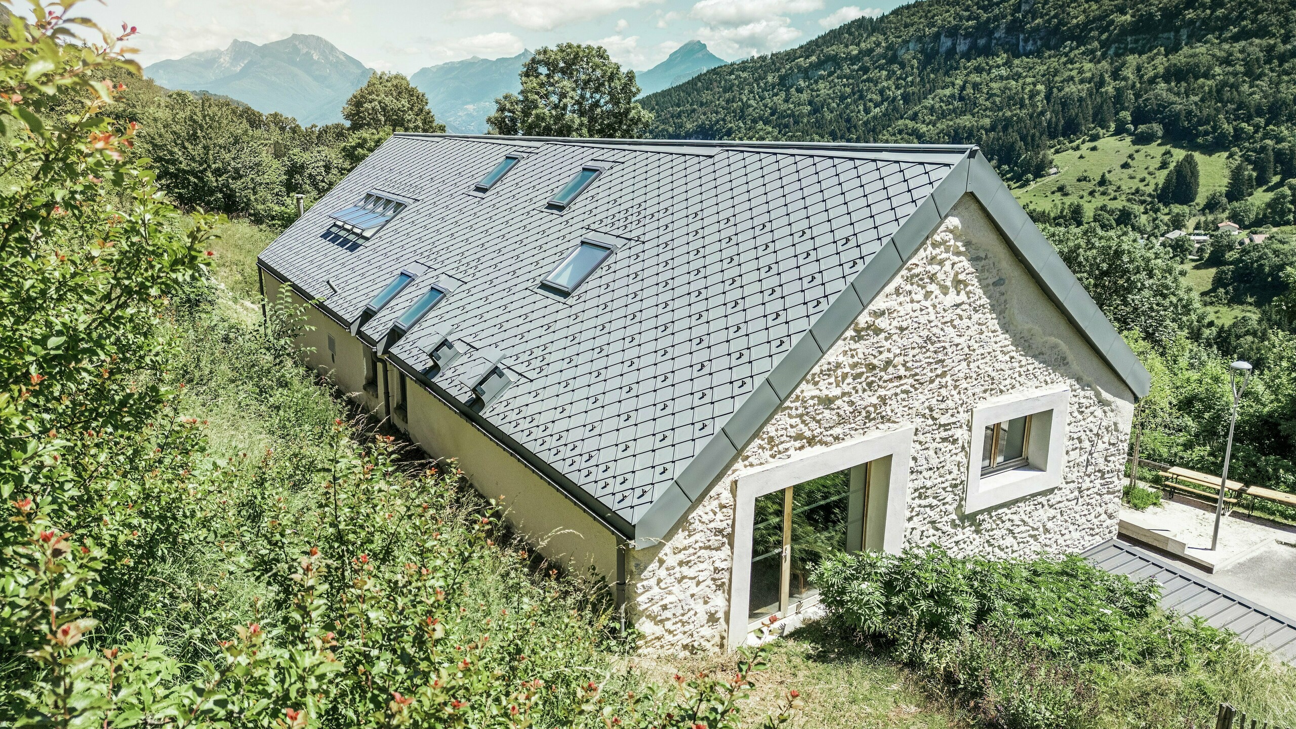 Blick auf das Dach der restaurierten Scheune „La Grange à Gaby“ in Engins, Frankreich. Das Gebäude ist mit langlebigen PREFA-Dachplatten im Format 29 × 29 cm gedeckt, die modernen Schutz mit traditioneller Ästhetik verbinden. Die idyllische Berglandschaft unterstreicht die harmonische Einbettung in die Natur.