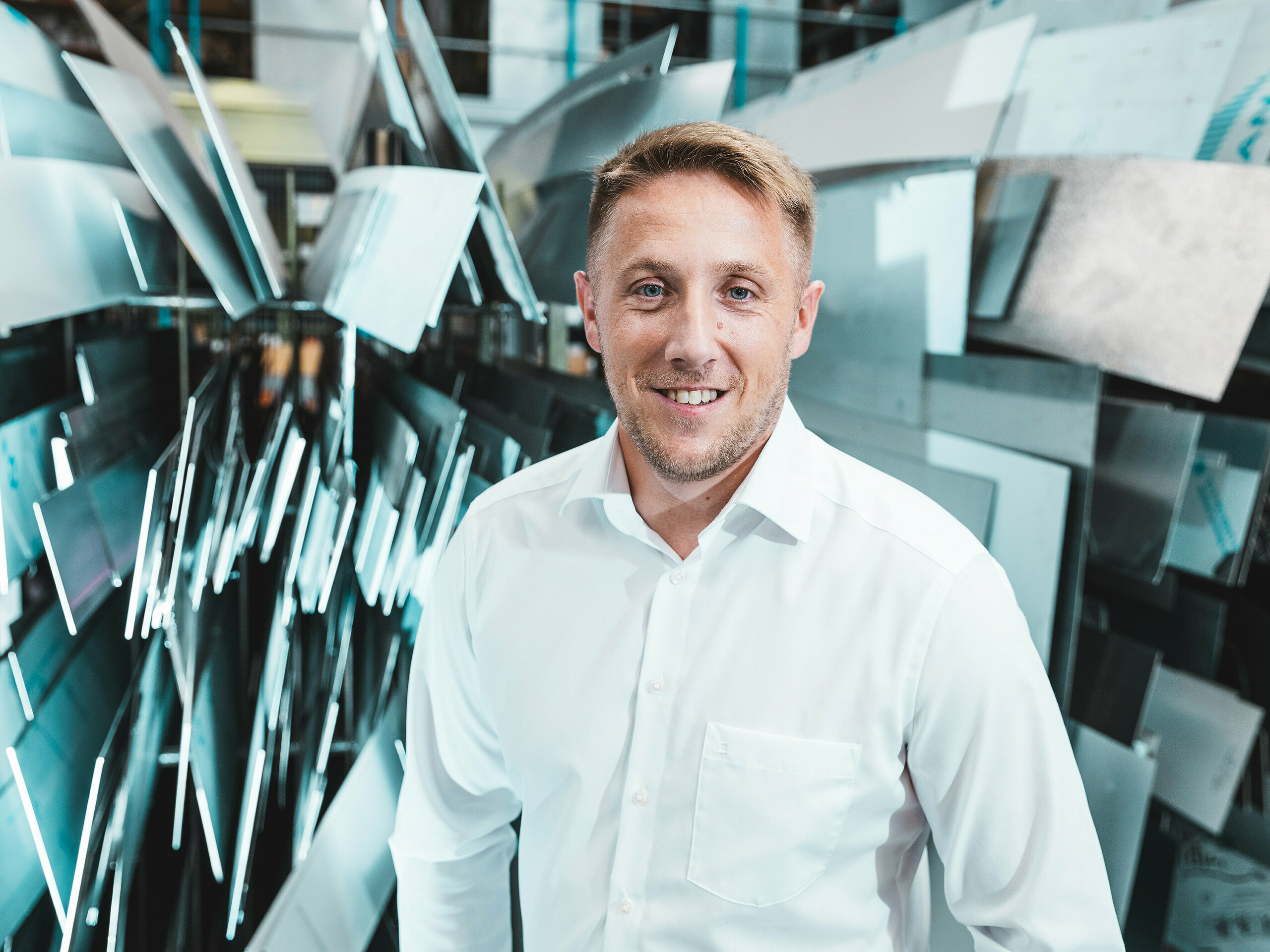 Projektový manažer Marcel Schmidt ze společnosti ASA Schüßler, která se zabývá kovovými střechami, stojí sebevědomě a přátelsky před působivou zdí zhotovenou z hliníkových prvků různých tvarů. Dynamicky uspořádané kovové panely efektivně odrážejí světlo a symbolizují inovaci, přesnost a kvalitu při zpracování kovů. Muž v bílé košili ztělesňuje kompetenci a moderní výrobní excelenci. V pozadí jsou patrné rozmanité možnosti zpracování hliníku – od střešních a fasádních systémů až po speciální řešení na míru.