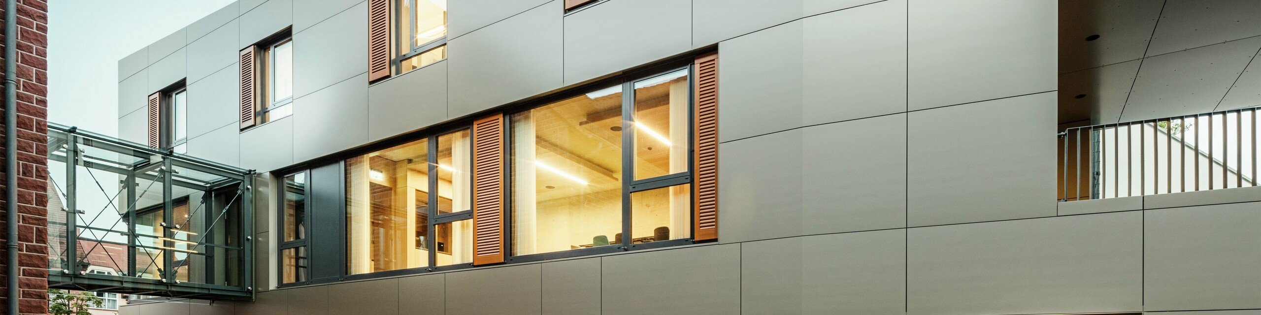 Moderní fasáda radnice v německém Alzenau je tvořena hliníkovými kompozitními panely PREFABOND v bronzové barvě, které jsou kombinovány s okenicemi a upravenými zelenými plochami. Vysoce kvalitní hliníkový obklad dodává elegantní a odolný vzhled a je ideální pro udržitelné stavební projekty. Boční strana je zde jasně viditelná.