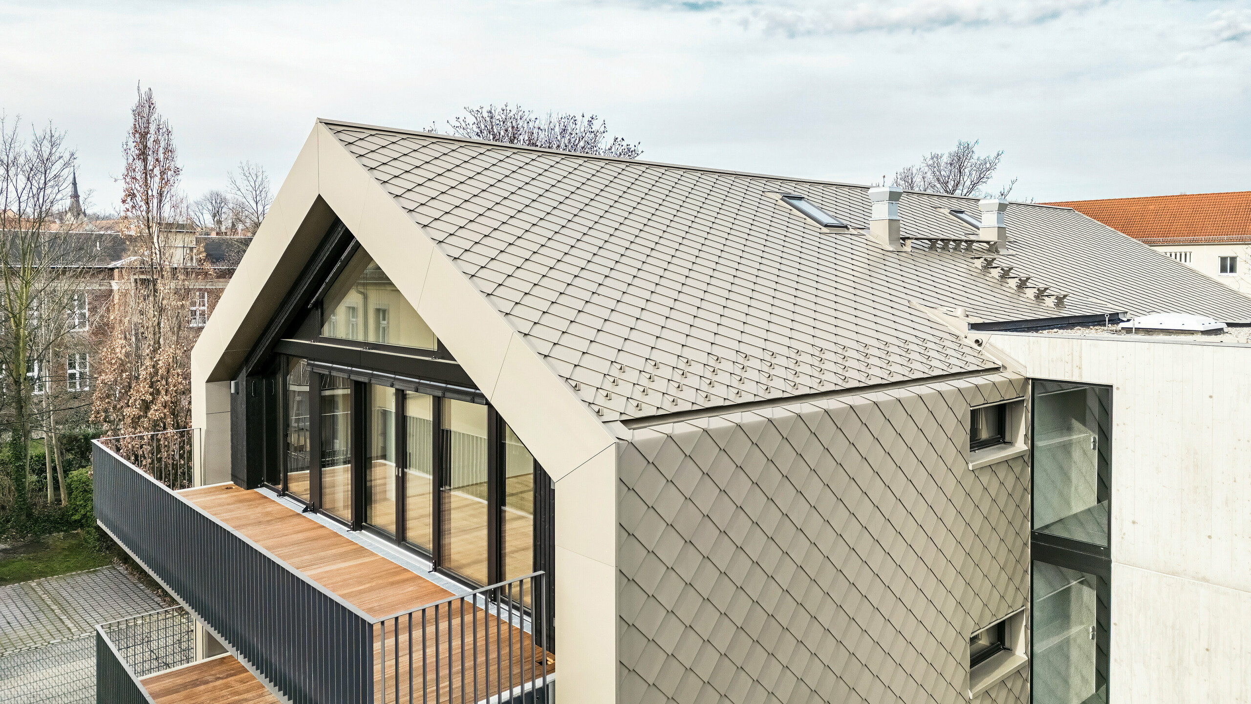 Pohled z ptačí perspektivy na moderní bytový dům na Chemnitzer Straße (Drážďany) s pláštěm falcovaných a fasádních šablon PREFA 29 × 29 v bronzu. Obrázek ukazuje elegantní střešní konstrukci, velká prosklená čela a balkony i harmonické začlenění budovy do okolí. Vysoce kvalitní materiály podtrhují dlouhodobou architekturu budovy.