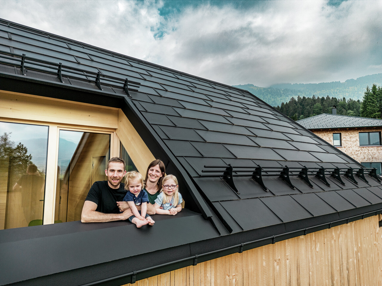 Die beiden Bauherren mit ihren Kindern genießen die Aussicht von der Loggia ihres neu erbauten Hauses, das mit stilvollen PREFA Solardachplatten in P.10 Schwarz ausgestattet ist.