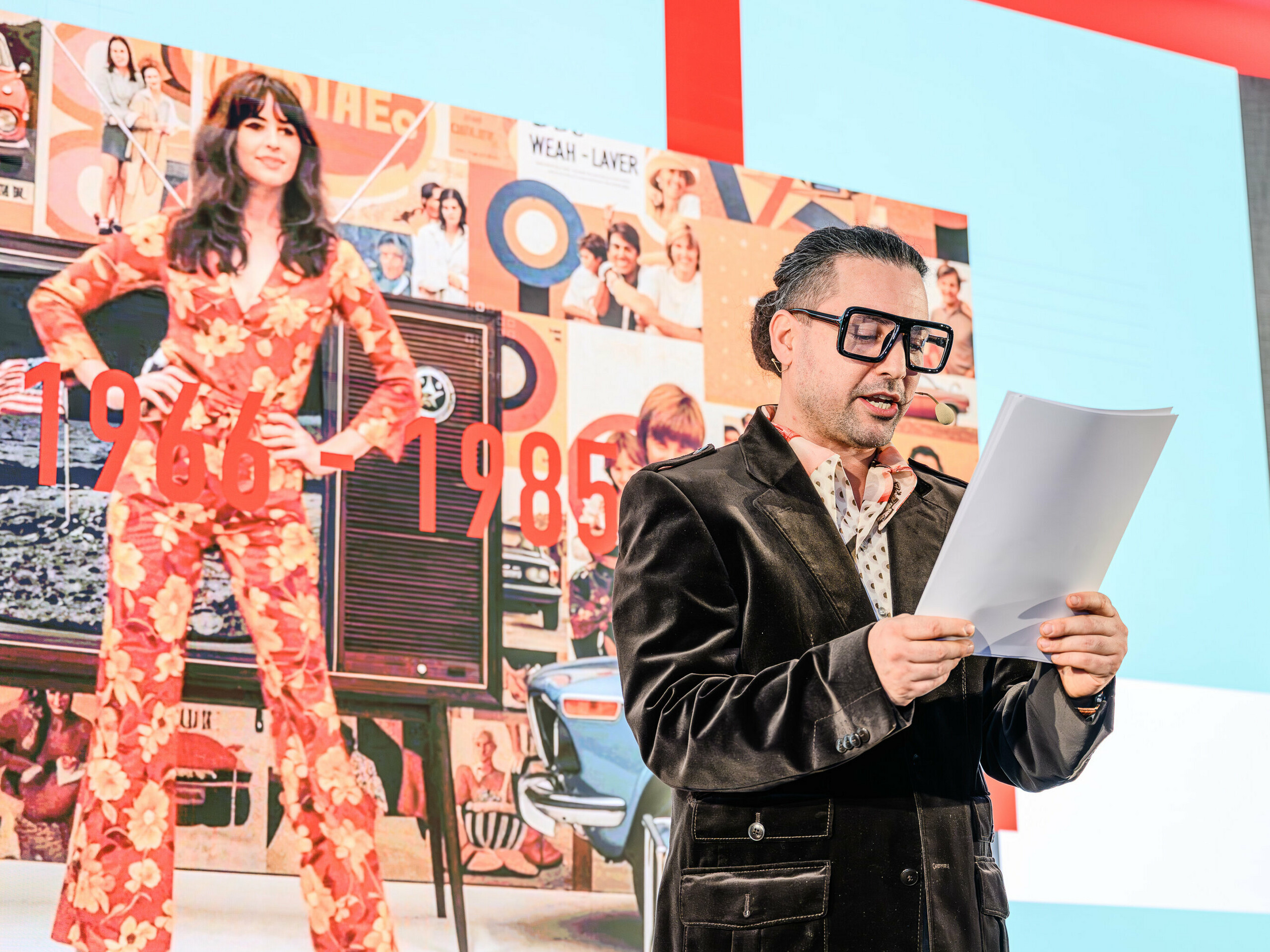 Cesta časem na pódiu na prezentaci PREFARENZEN 2026: Charismatický Serkan Temel v retro vzhledu provede diváky desetiletími. Důraz je kladen na roky 1966 až 1985, zobrazené v koláži v pozadí s ikonami designu a společenskými trendy dané doby. Stejně jako produkty PREFA, i trendy přetrvávají stylově a obsahově. Prezentace ukazuje, jak architektura včerejška inspiruje zítřejší řešení – reinterpretovaná hliníkem.